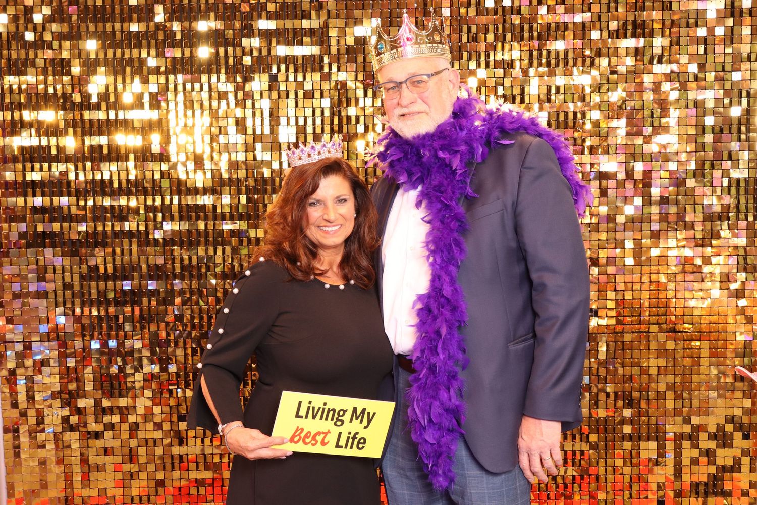 Couple poses with props including a crown and purple boa in front of a gold sequin backdrop.