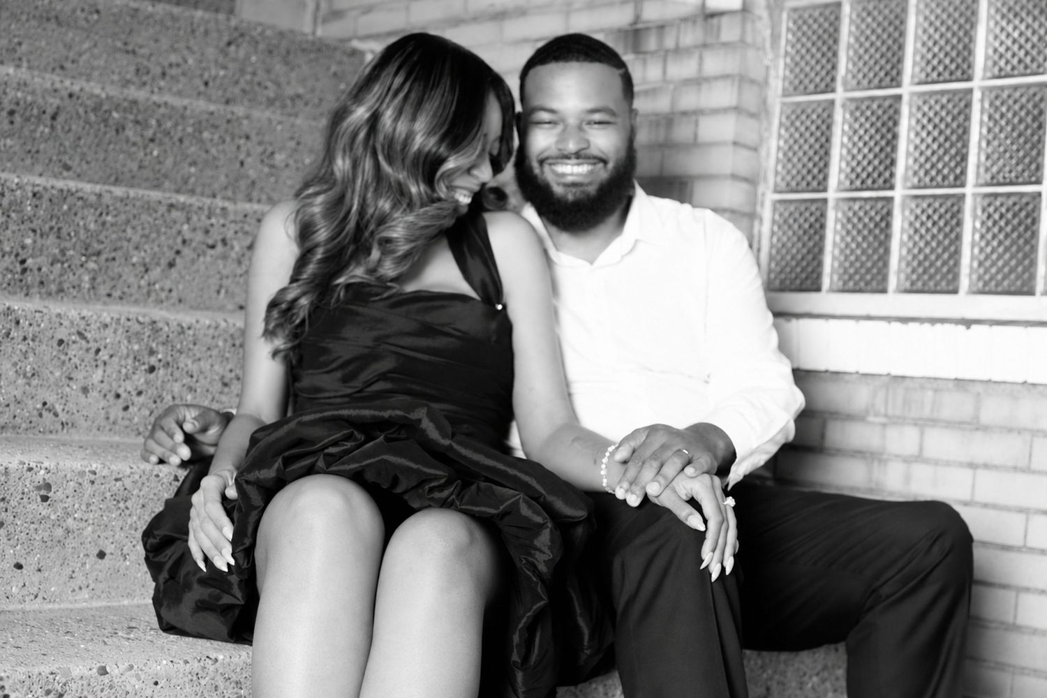 Black and white photo of a smiling couple sitting on steps, woman in black dress and man in white shirt.