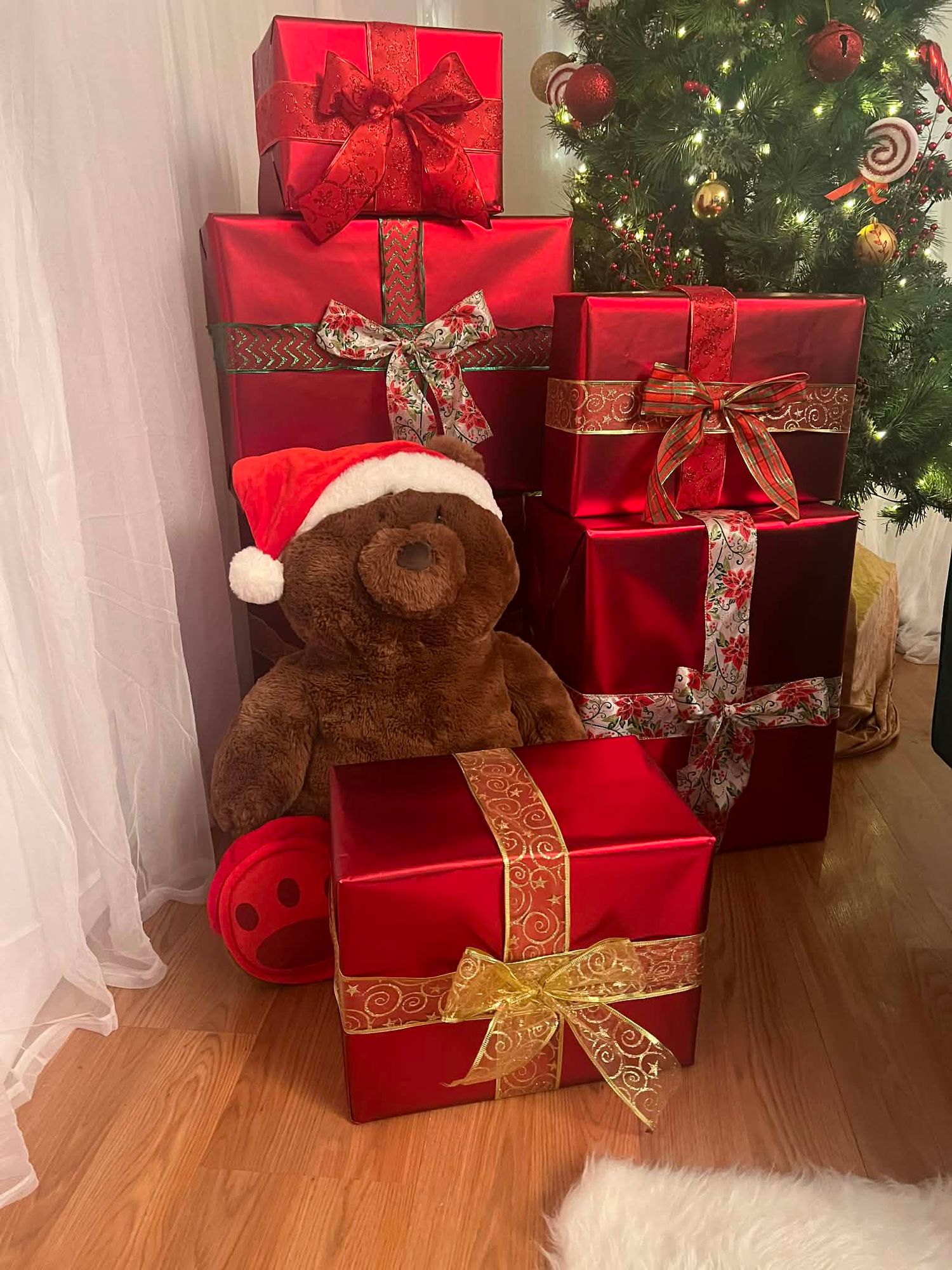 A brown teddy bear wearing a Santa hat sits among stacked red Christmas gift boxes by a tree.
