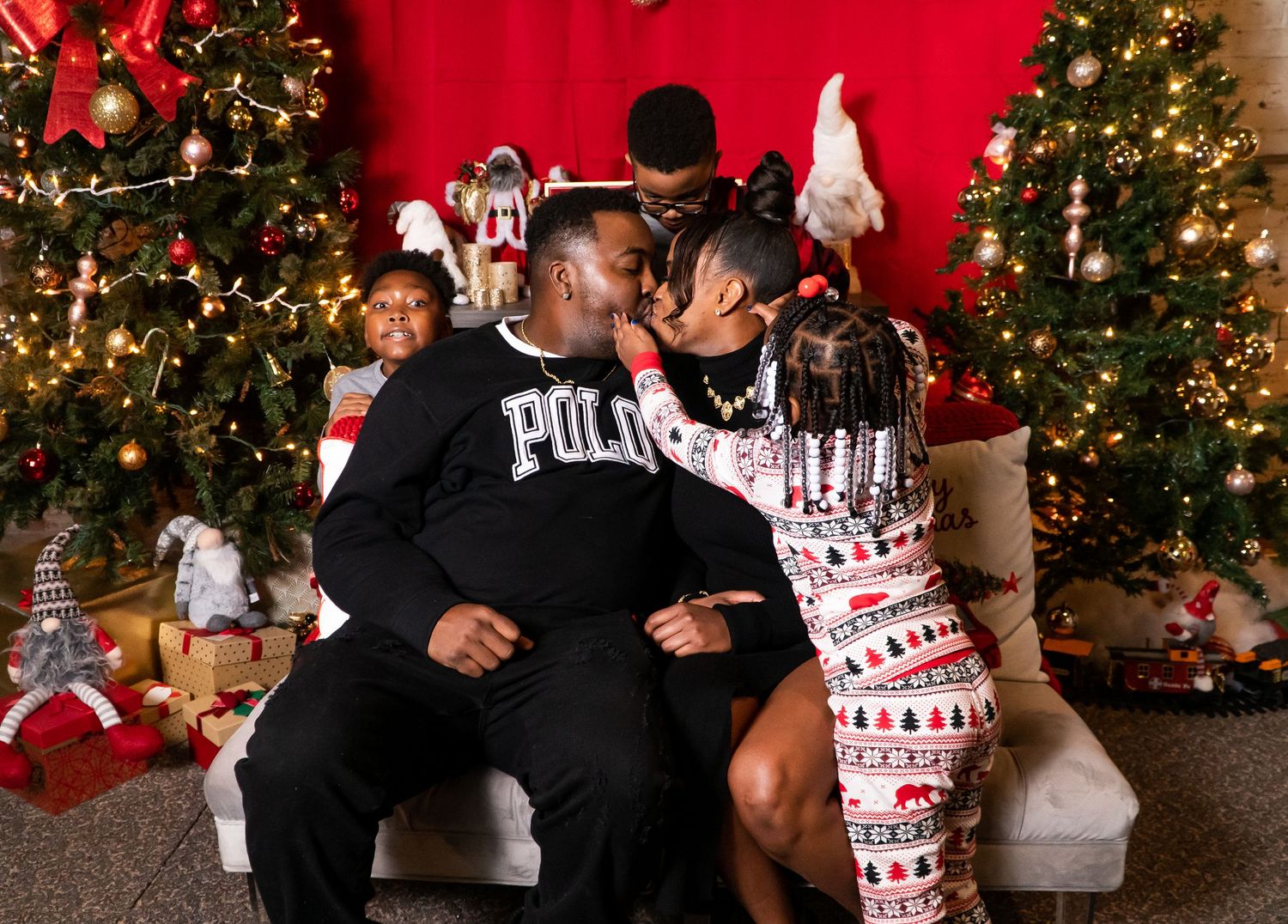 Couple sharing a kiss during a festive Christmas photo session with decorated trees and gifts.