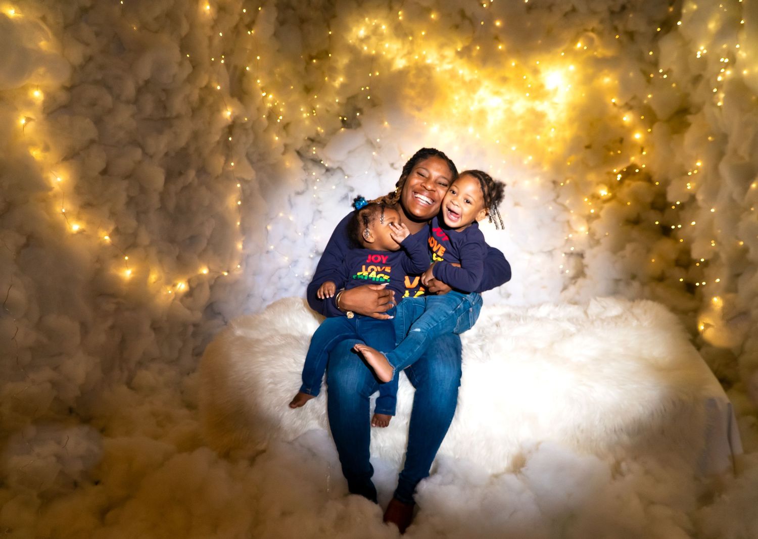 Mother embracing two young children surrounded by glowing fairy lights in dreamy setting.