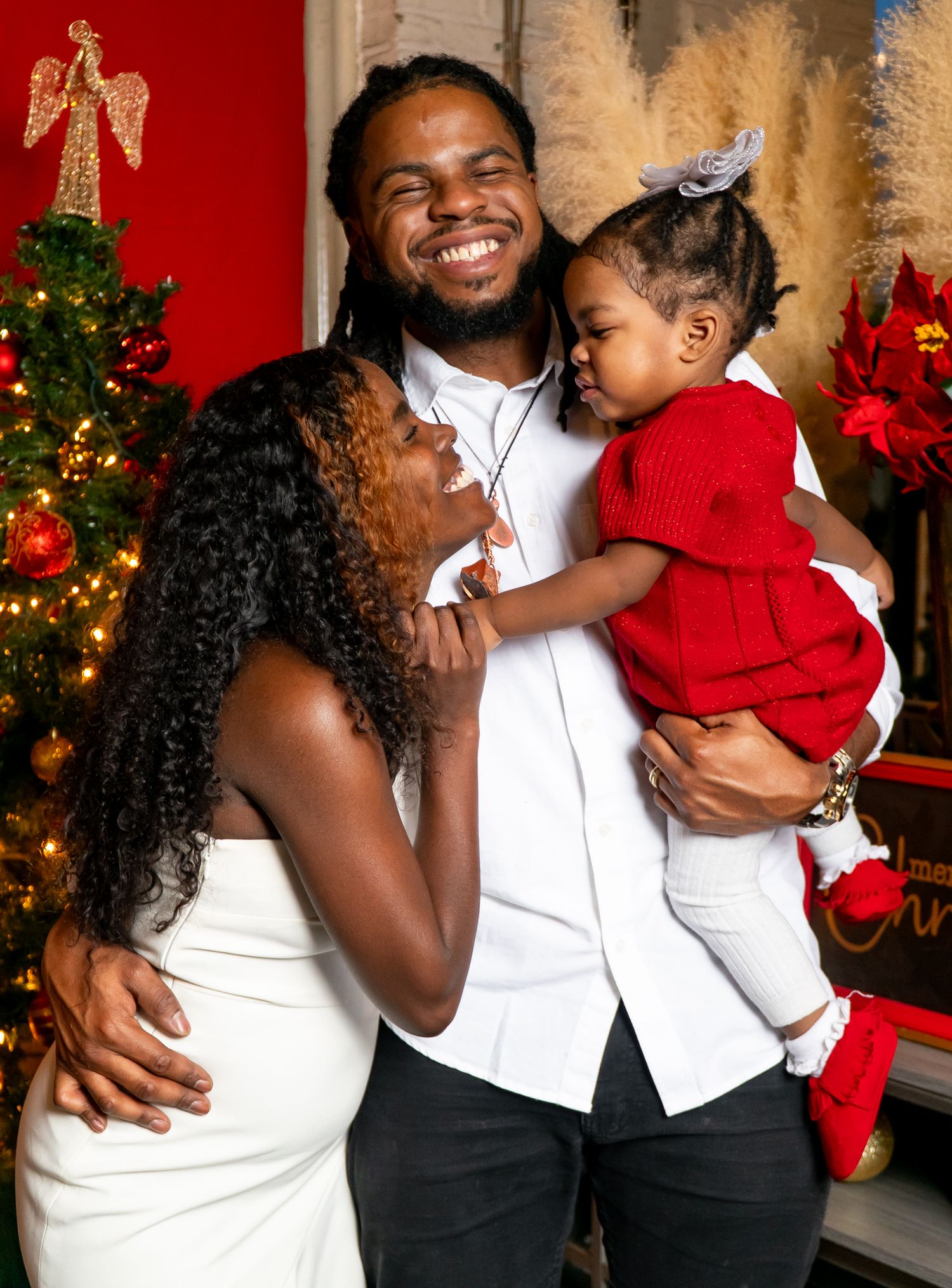 Smiling man holds a baby in red while a woman leans in during a Christmas photo session.