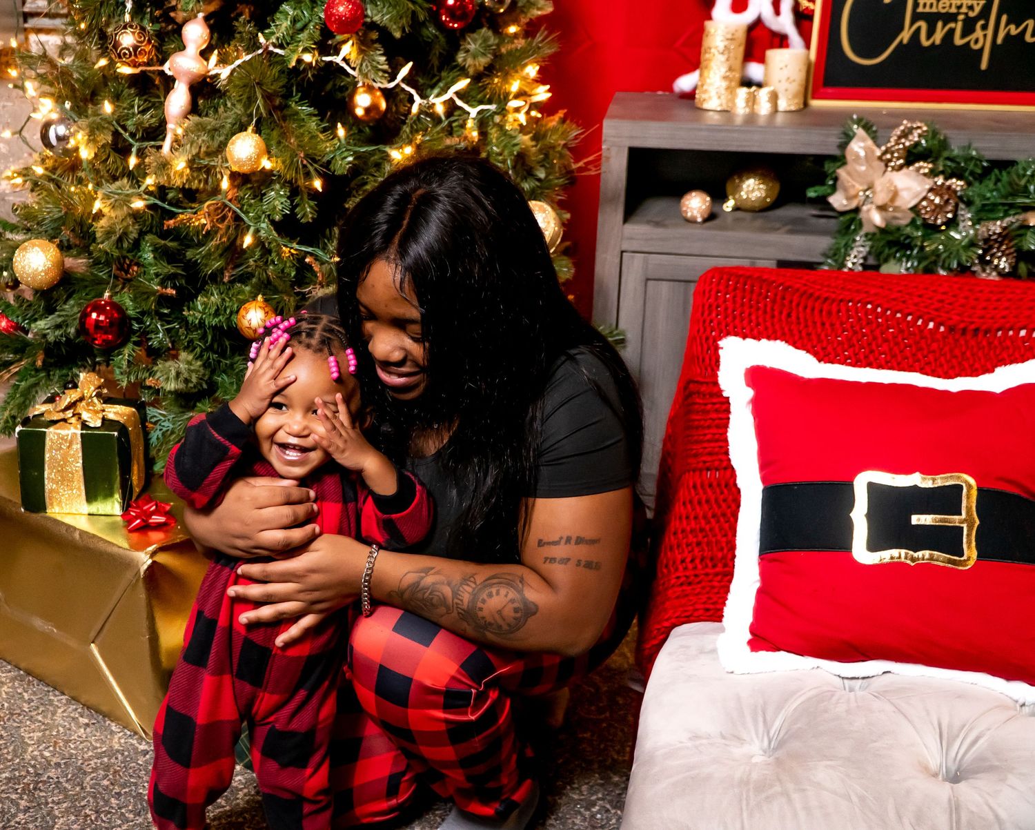 Mother hugging laughing toddler in matching red plaid pajamas beside decorated Christmas tree.