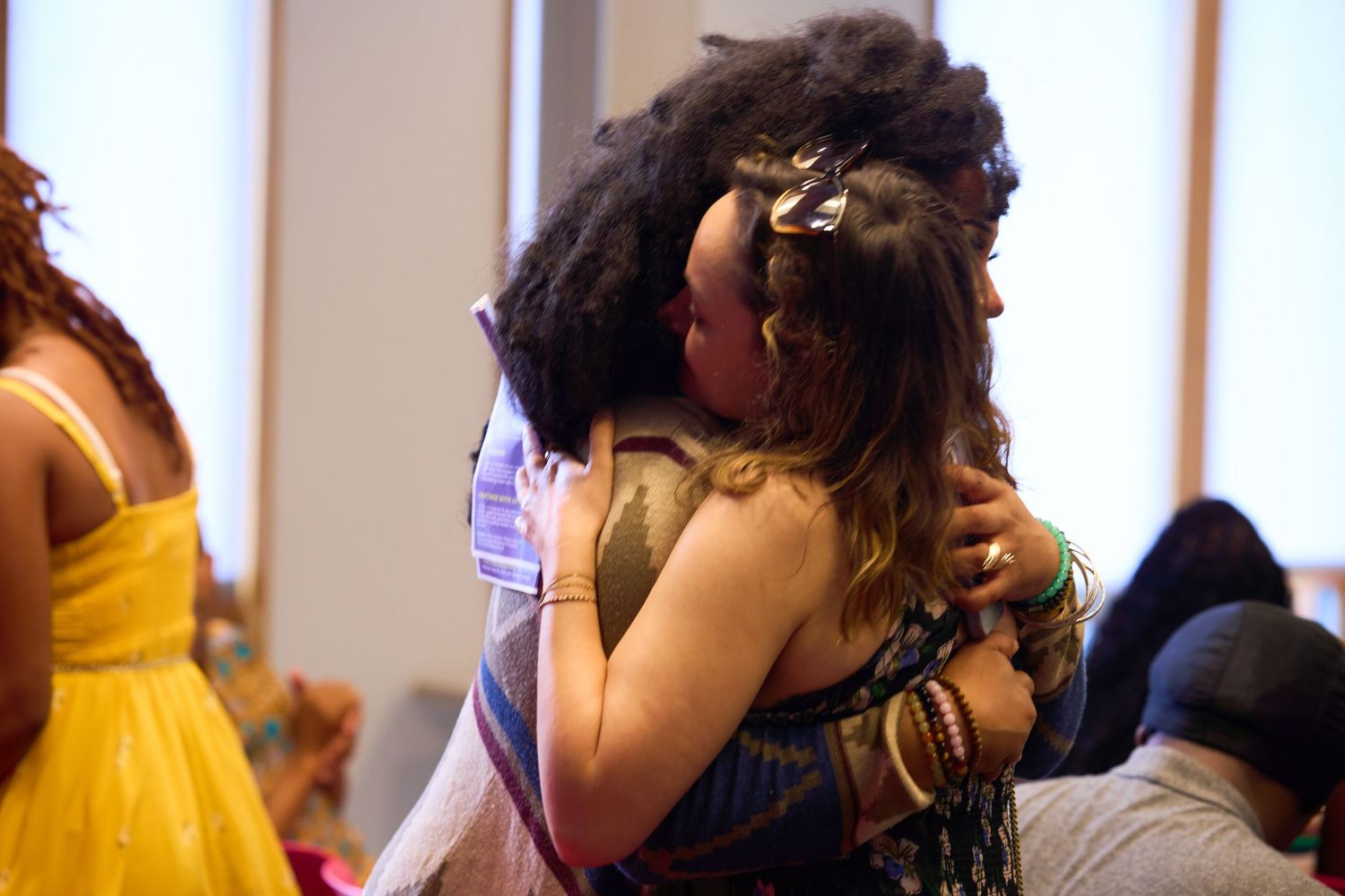 Two women sharing a warm embrace at an indoor event, with guests visible in the background.