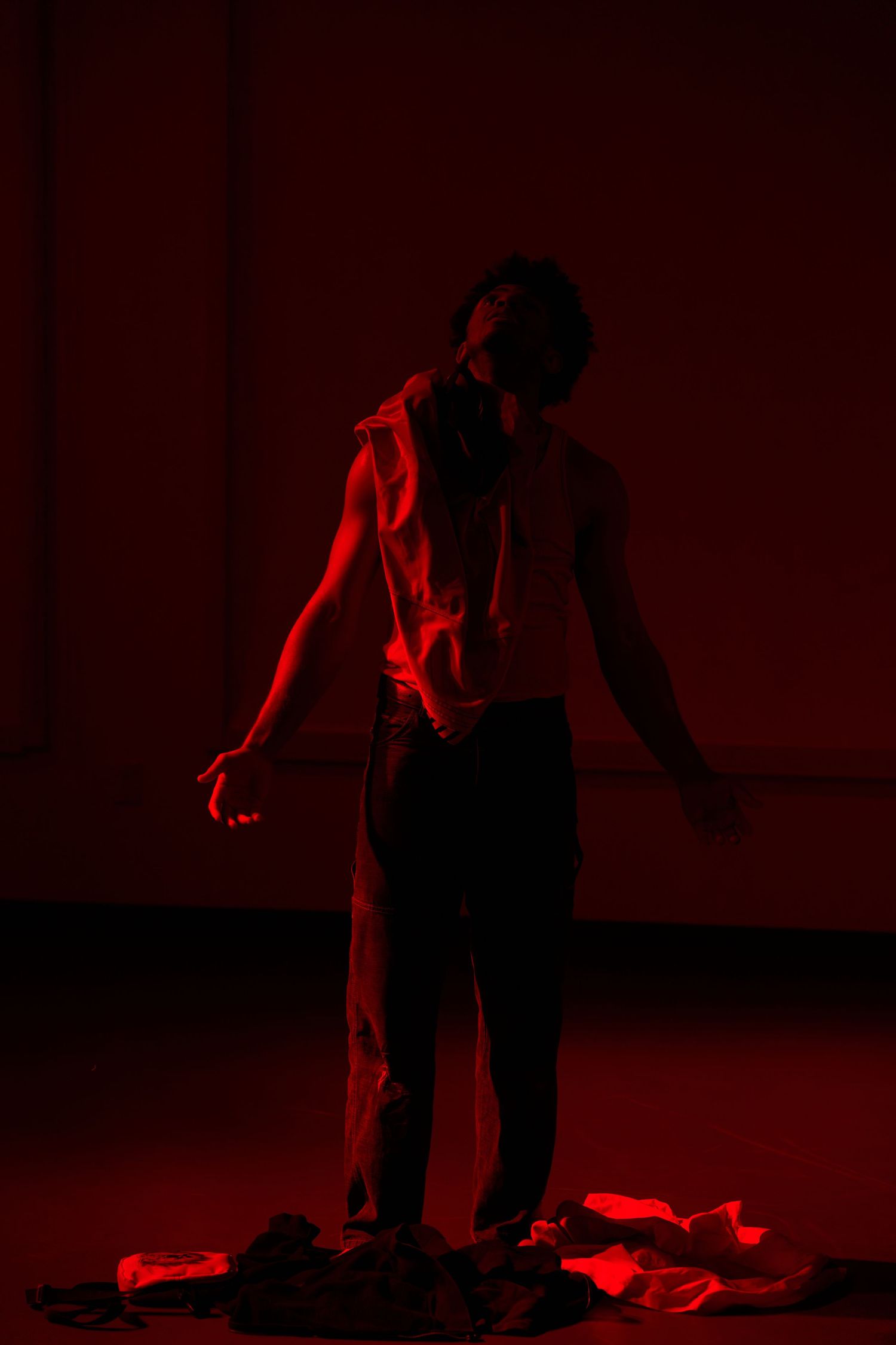 A performer stands dramatically under red stage lighting, wearing an open vest, with fabric scattered on the floor.