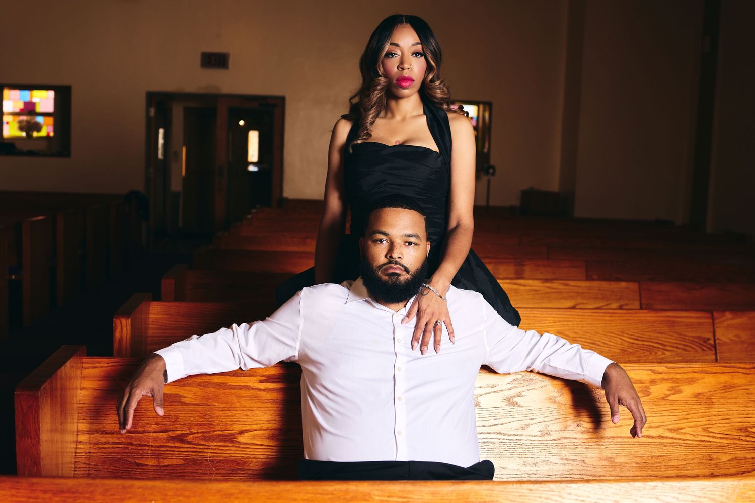 A couple poses in a church, man in white shirt sits in pew while woman in black dress stands behind him.