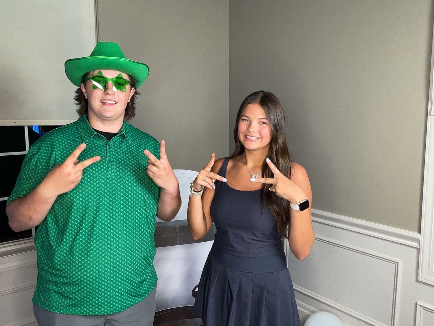 Two friends pose making hand signs, one wearing a green St. Patrick's Day hat and shirt, the other in a black dress.