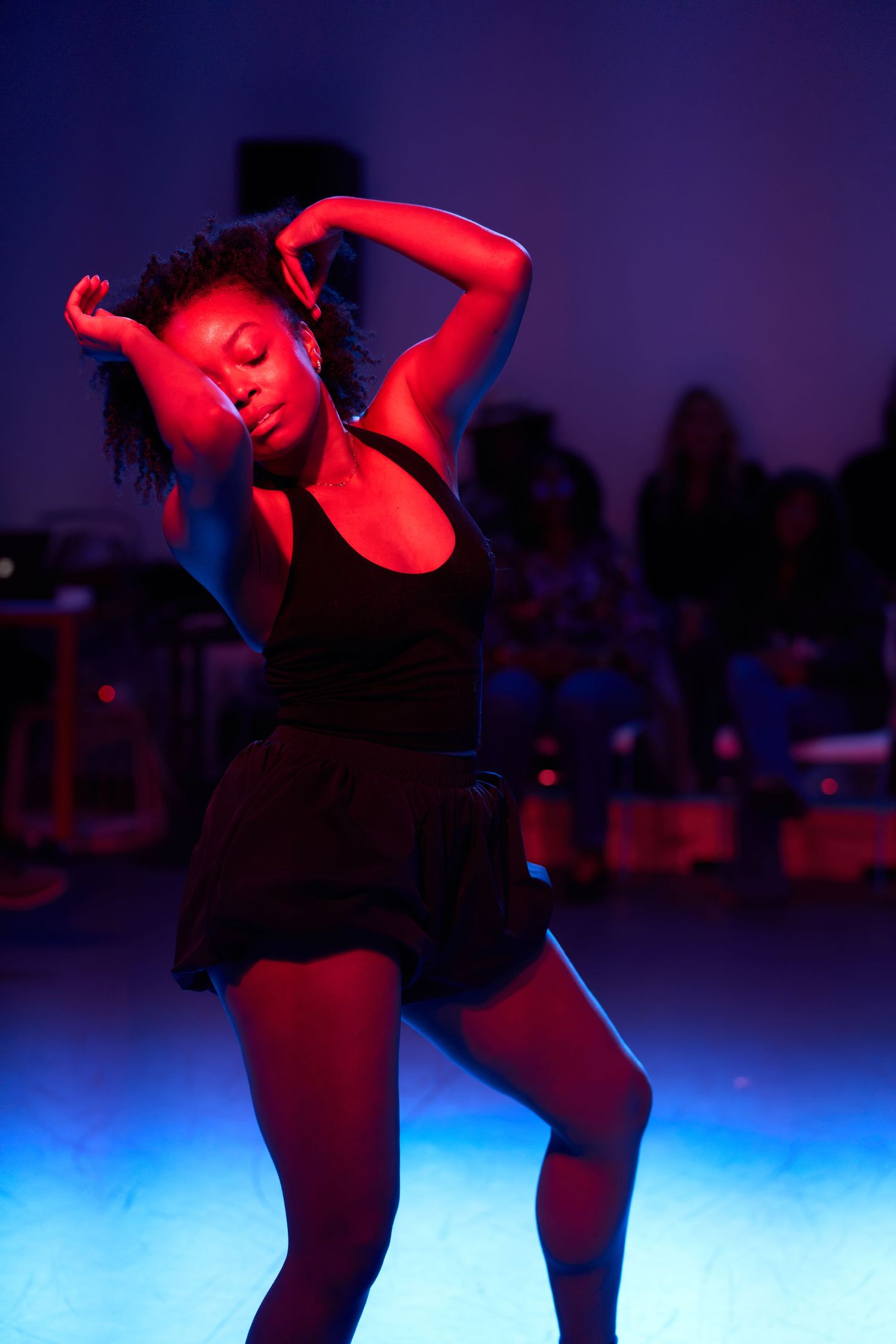 A dancer performs under dramatic red and blue stage lighting, wearing black outfit with eyes closed and arms raised.