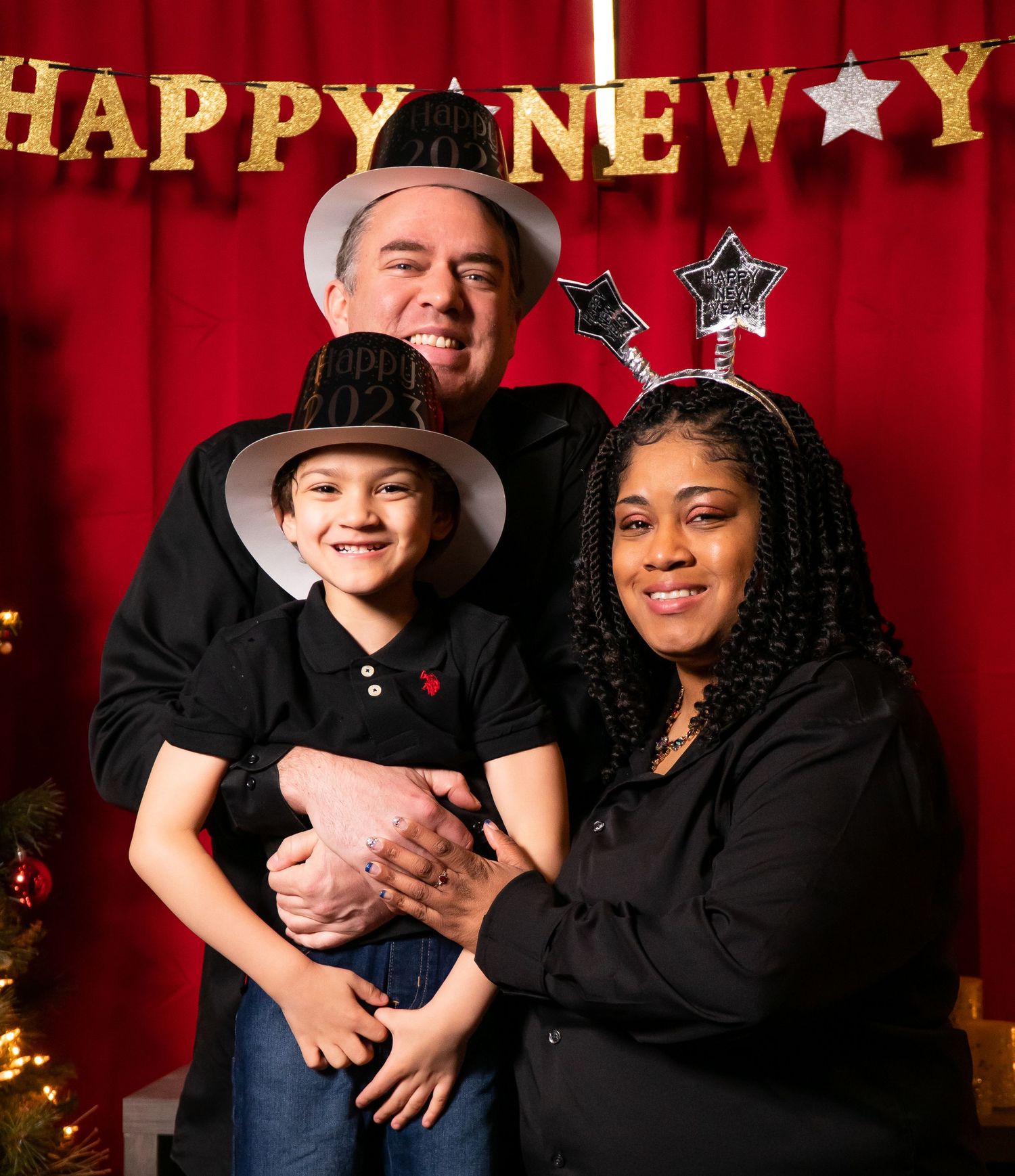 Family of three celebrates New Year wearing festive hats under a glittery Happy New Year banner.