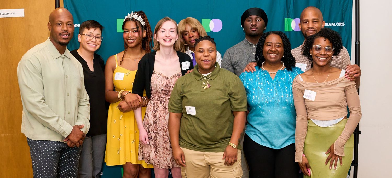 Diverse group of smiling adults posing together at a community event in front of a teal backdrop.