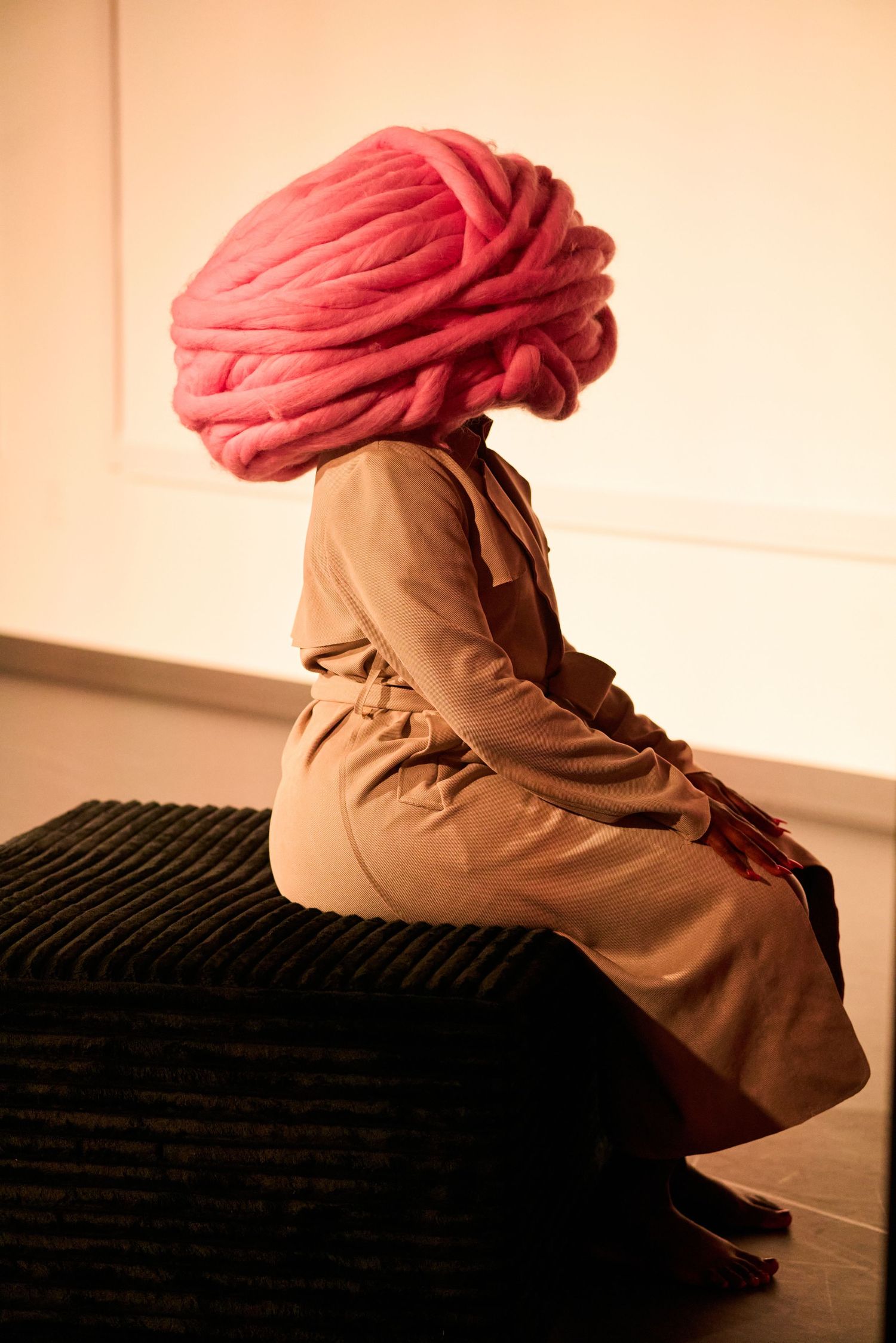Person sitting on black ottoman wearing tan coat with large pink yarn turban covering their head in gallery.