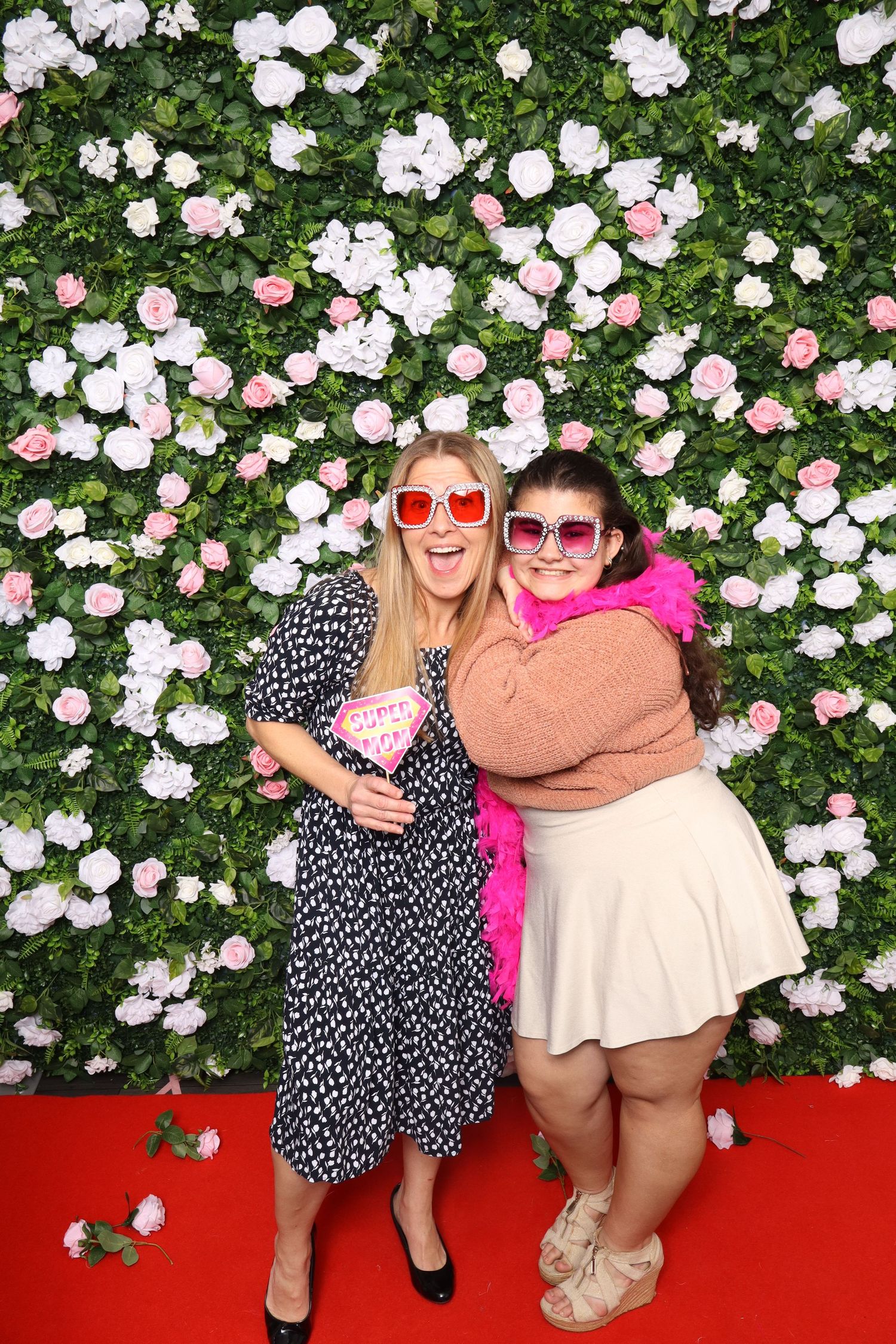 Two smiling women wearing heart-shaped sunglasses pose in front of a white and pink floral wall backdrop.