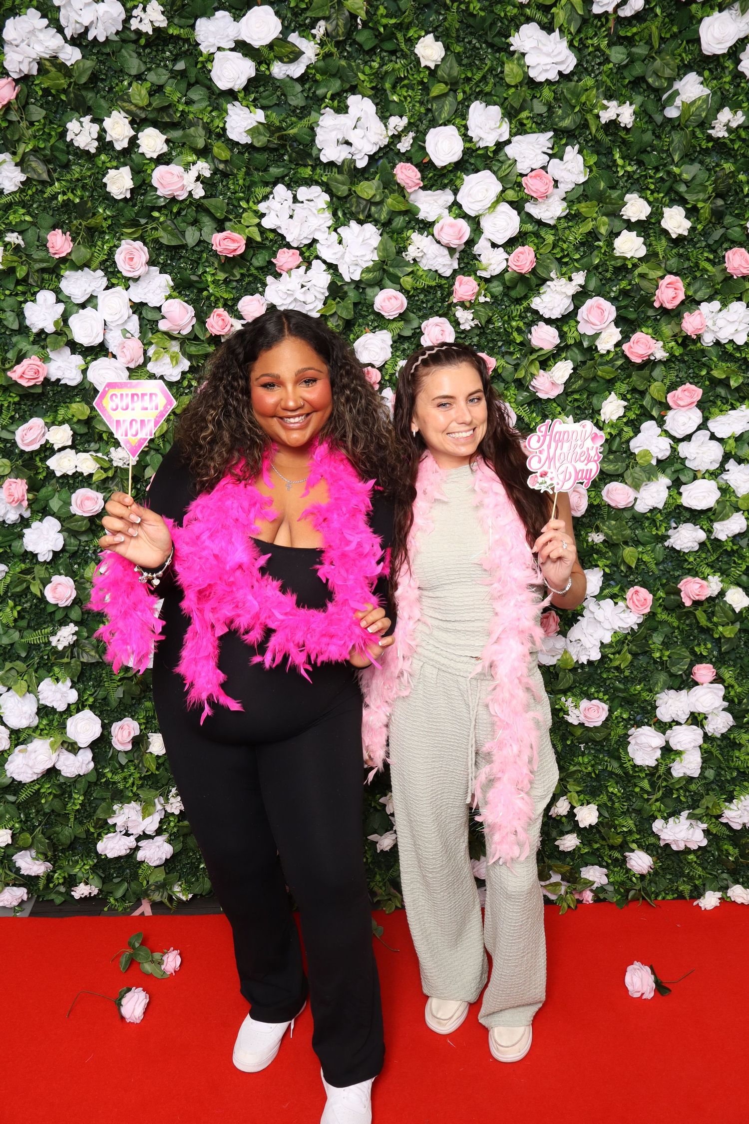 Two women pose with pink feather boas and props in front of a floral wall backdrop on a red carpet.