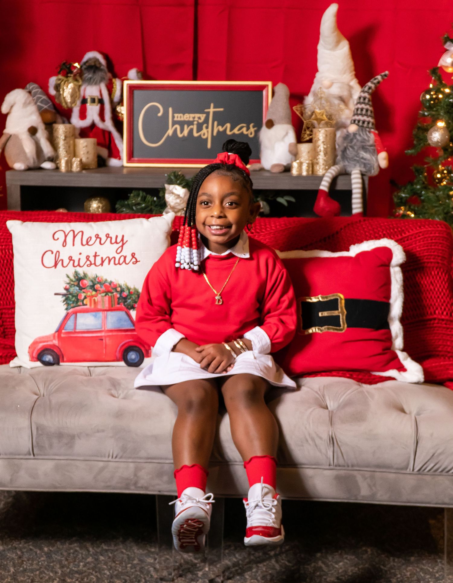 Young girl in red Christmas outfit sitting on a couch surrounded by holiday decorations.