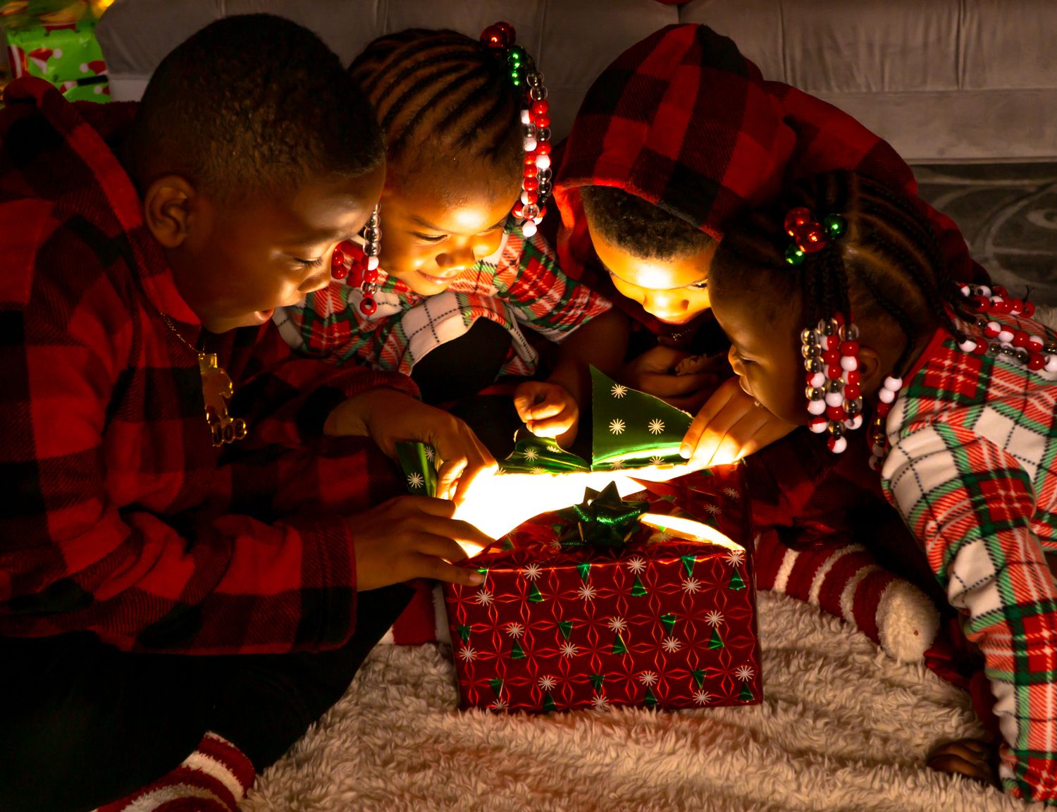 Children in plaid pajamas opening glowing Christmas gift together by firelight.
