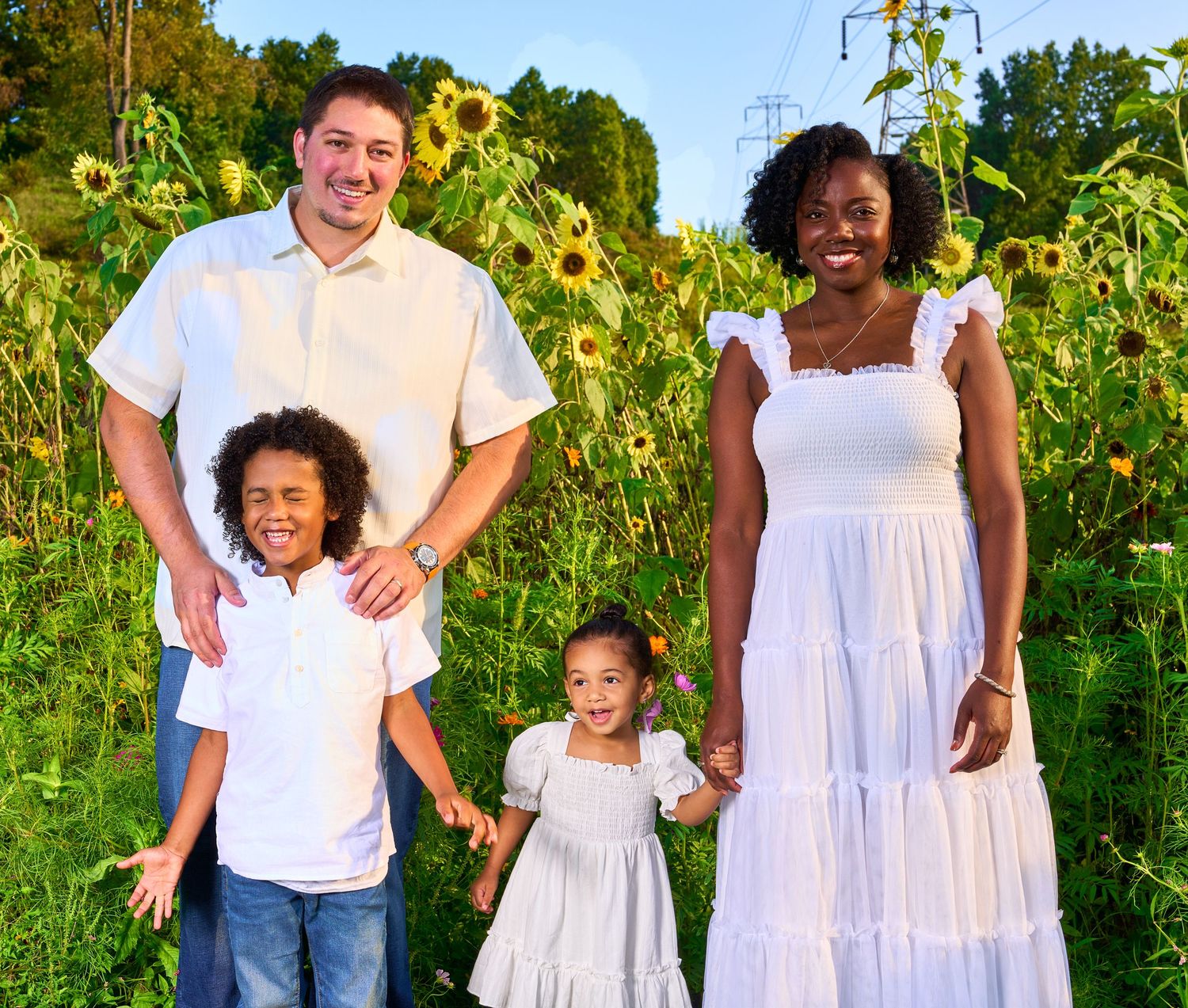 Happy interracial family of four posing in a sunflower field, parents and two young children wearing white outfits.