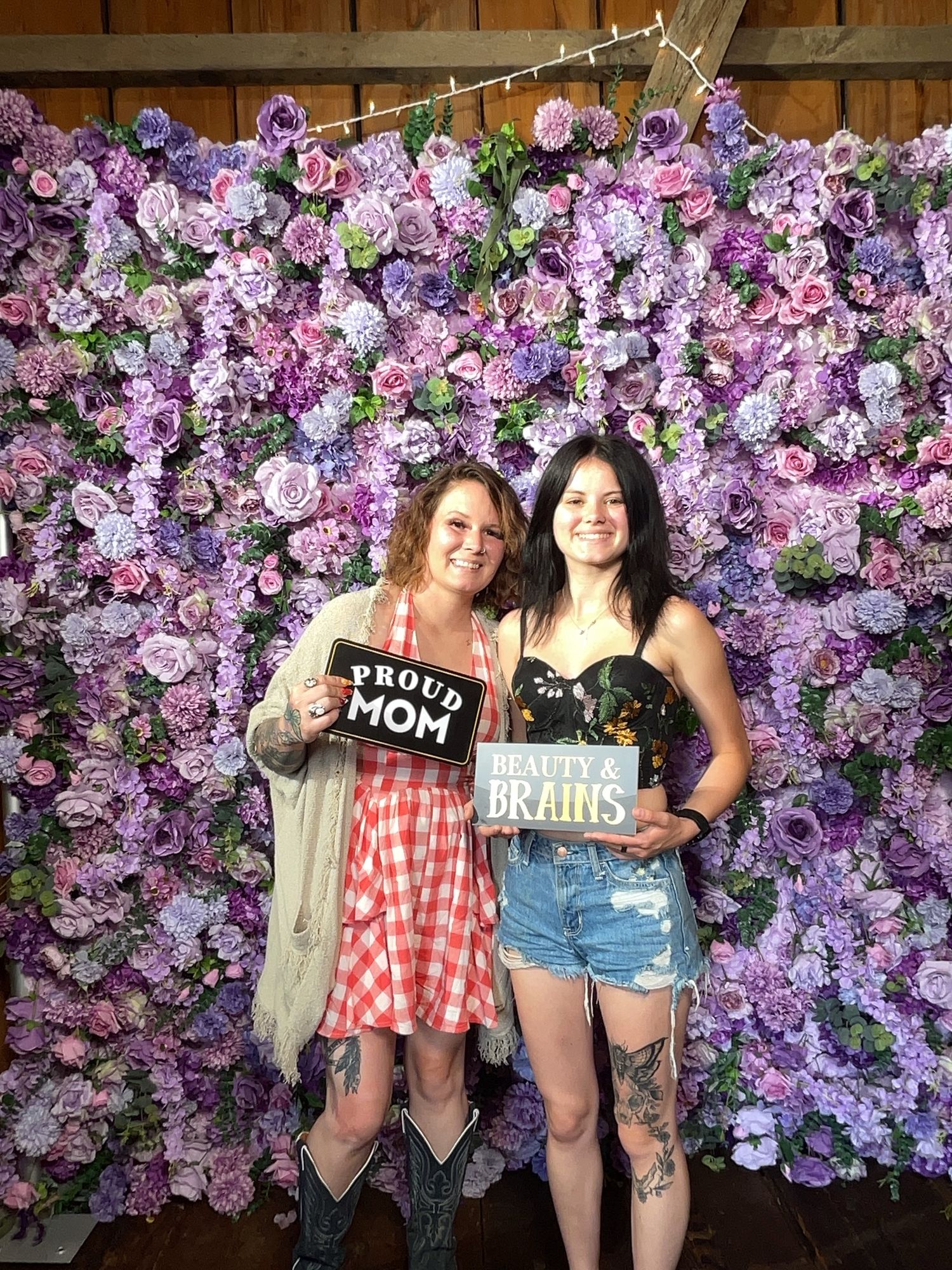 Two women pose in front of a purple floral wall, holding signs reading Proud Mom and Beauty & Brains.