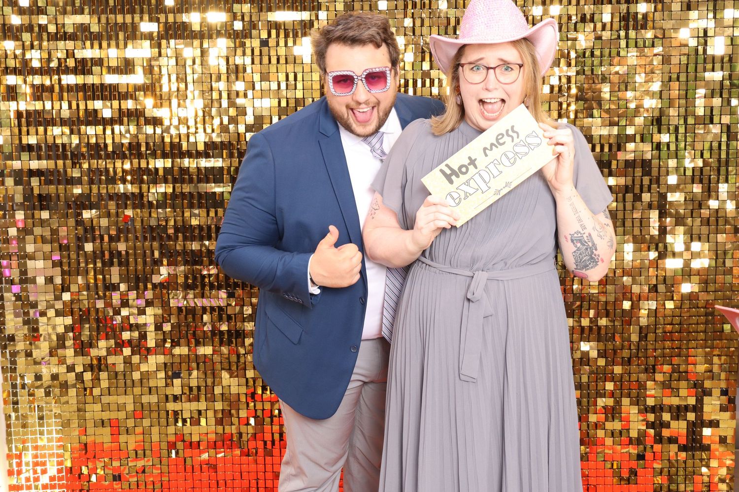Two people posing in front of a gold sequin backdrop, man in blue suit and woman in gray dress holding a sign.