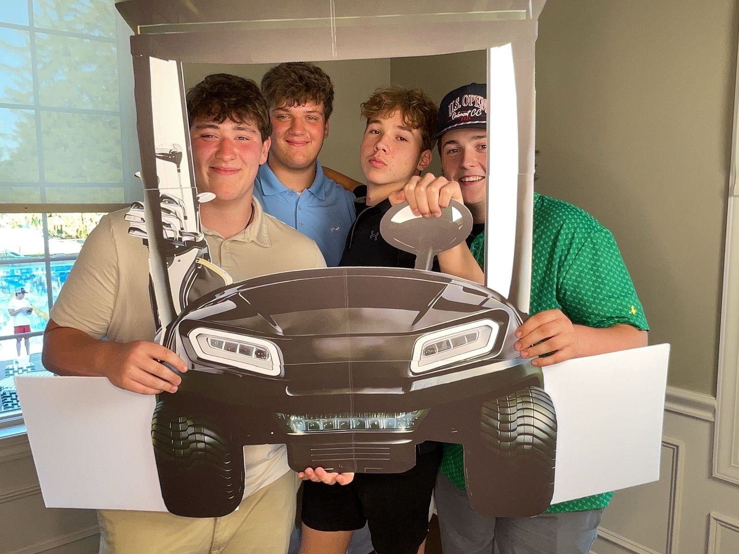 Four teenage boys posing with a cardboard car cutout prop for a fun photo indoors.