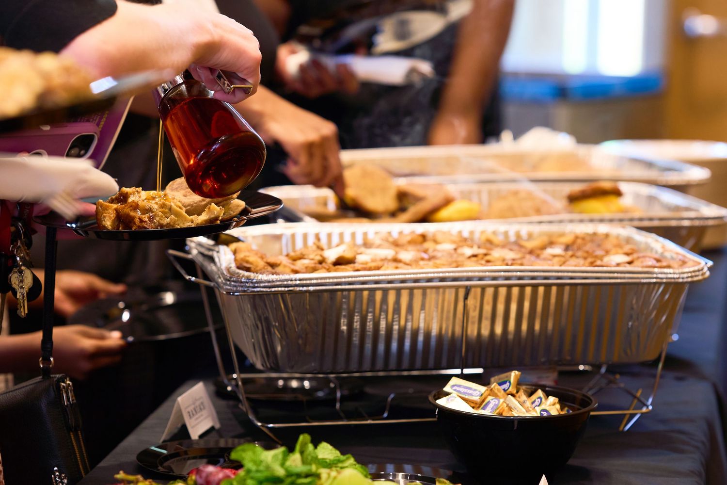 Buffet table with chafing dishes filled with food, as a person pours syrup over a dish at a catered event.