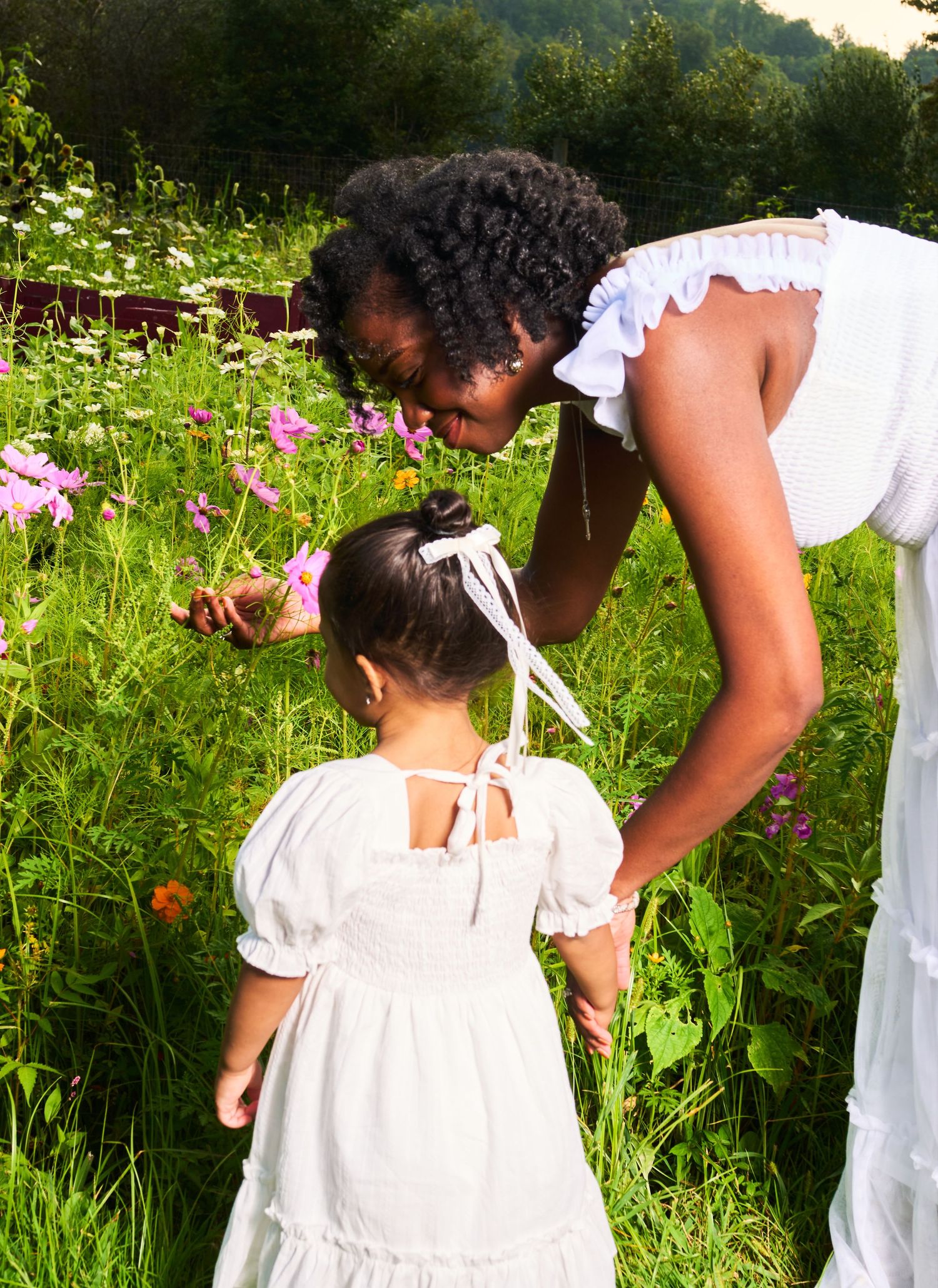 A woman and young girl in white dresses explore wildflowers in a lush green meadow on a sunny day.