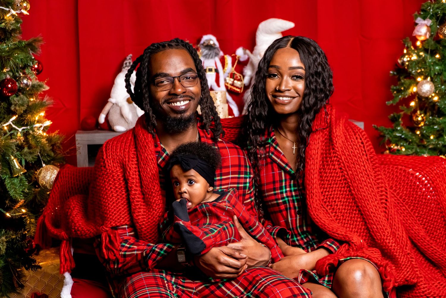 Family of three in matching red plaid outfits posing together by Christmas tree with Santa.