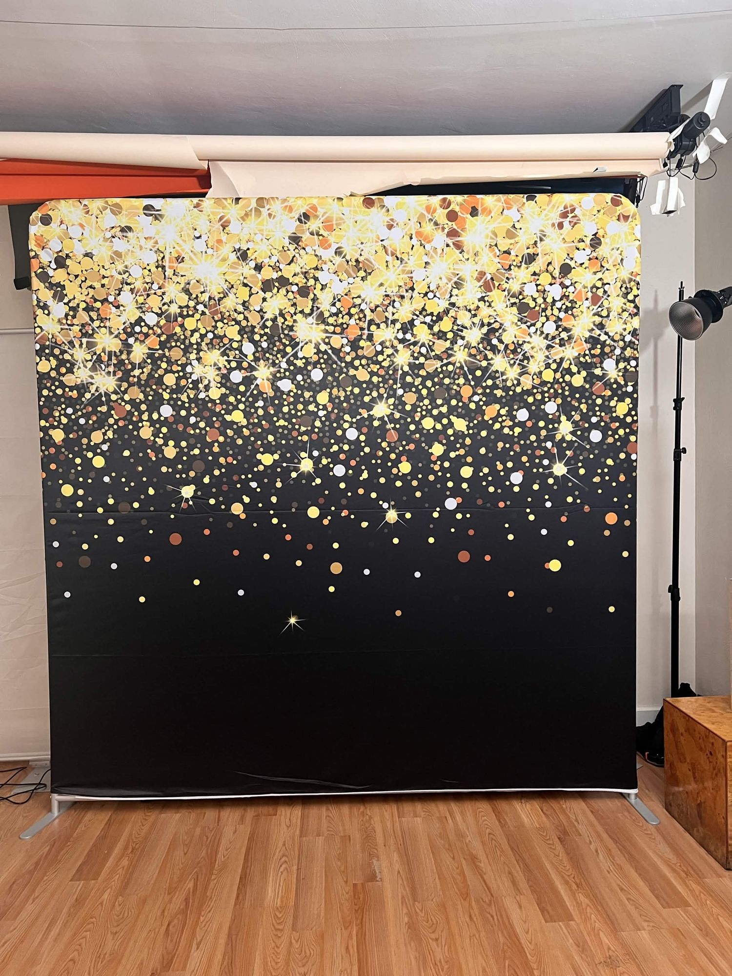 Black and gold confetti glitter photo booth backdrop on a metal stand in a photography studio.