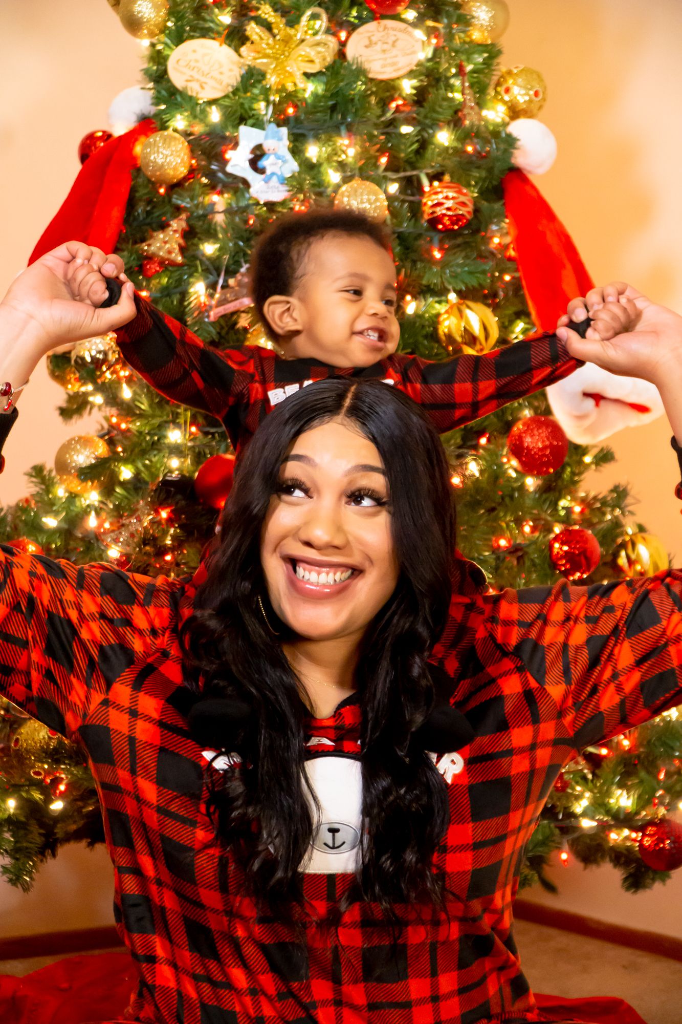 Smiling mother with toddler on shoulders in matching red plaid pajamas by Christmas tree.