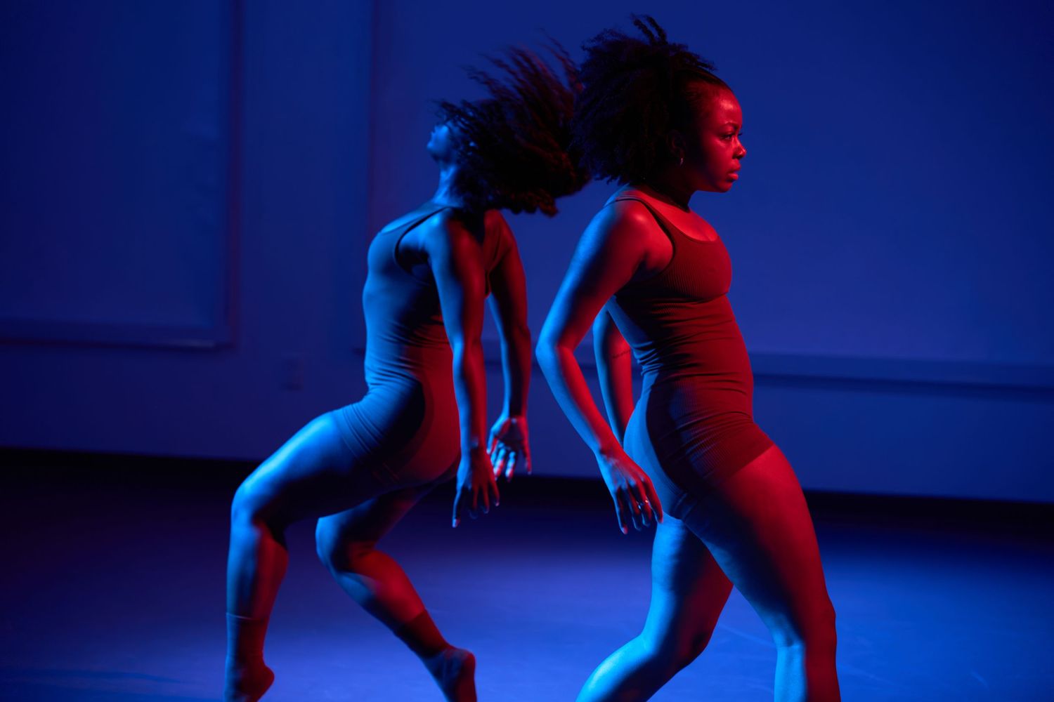 Two female dancers perform under dramatic red and blue stage lighting in a contemporary dance performance.