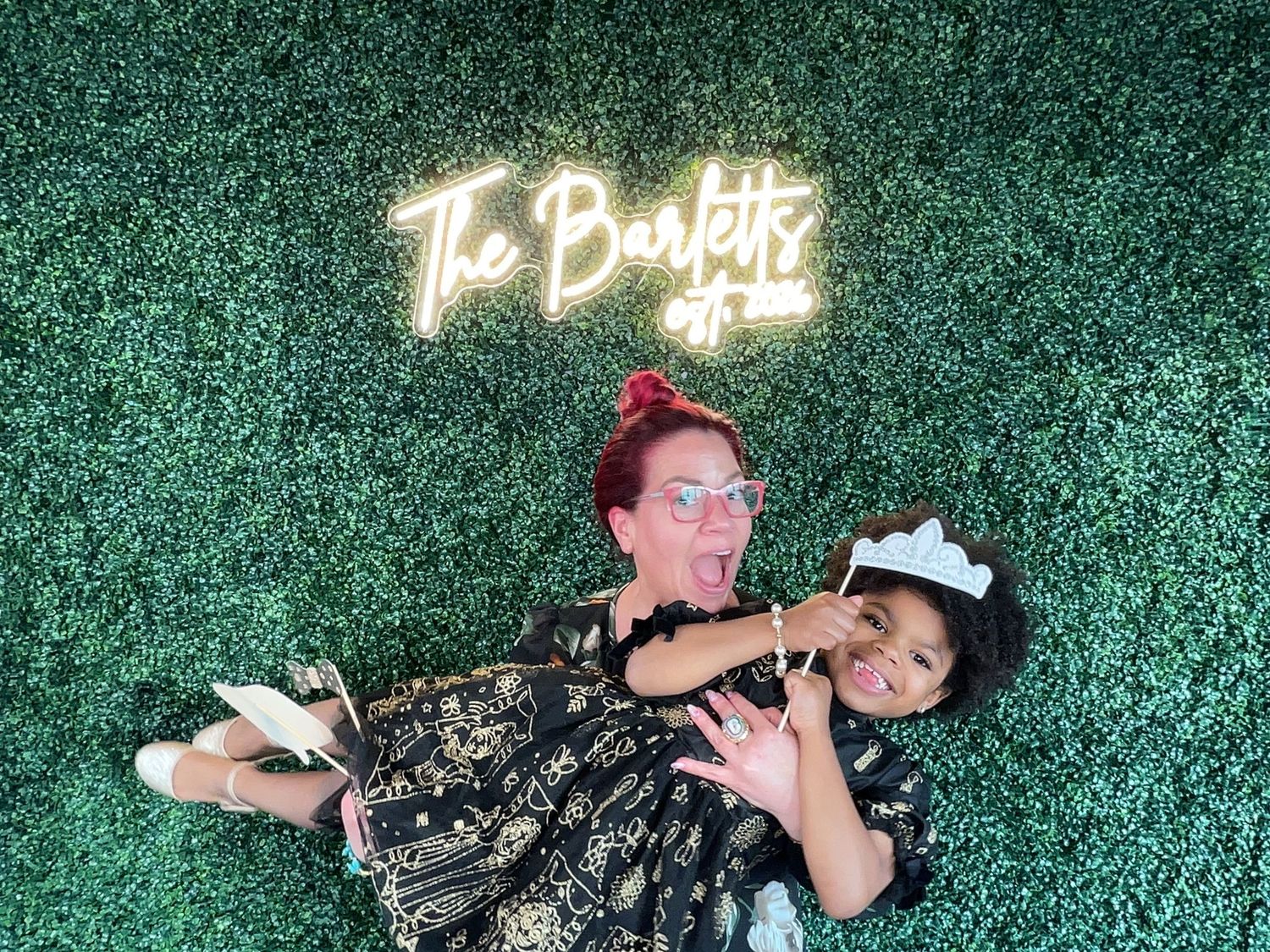 Two women pose playfully in front of a green hedge wall with a neon sign reading The Bartetts.