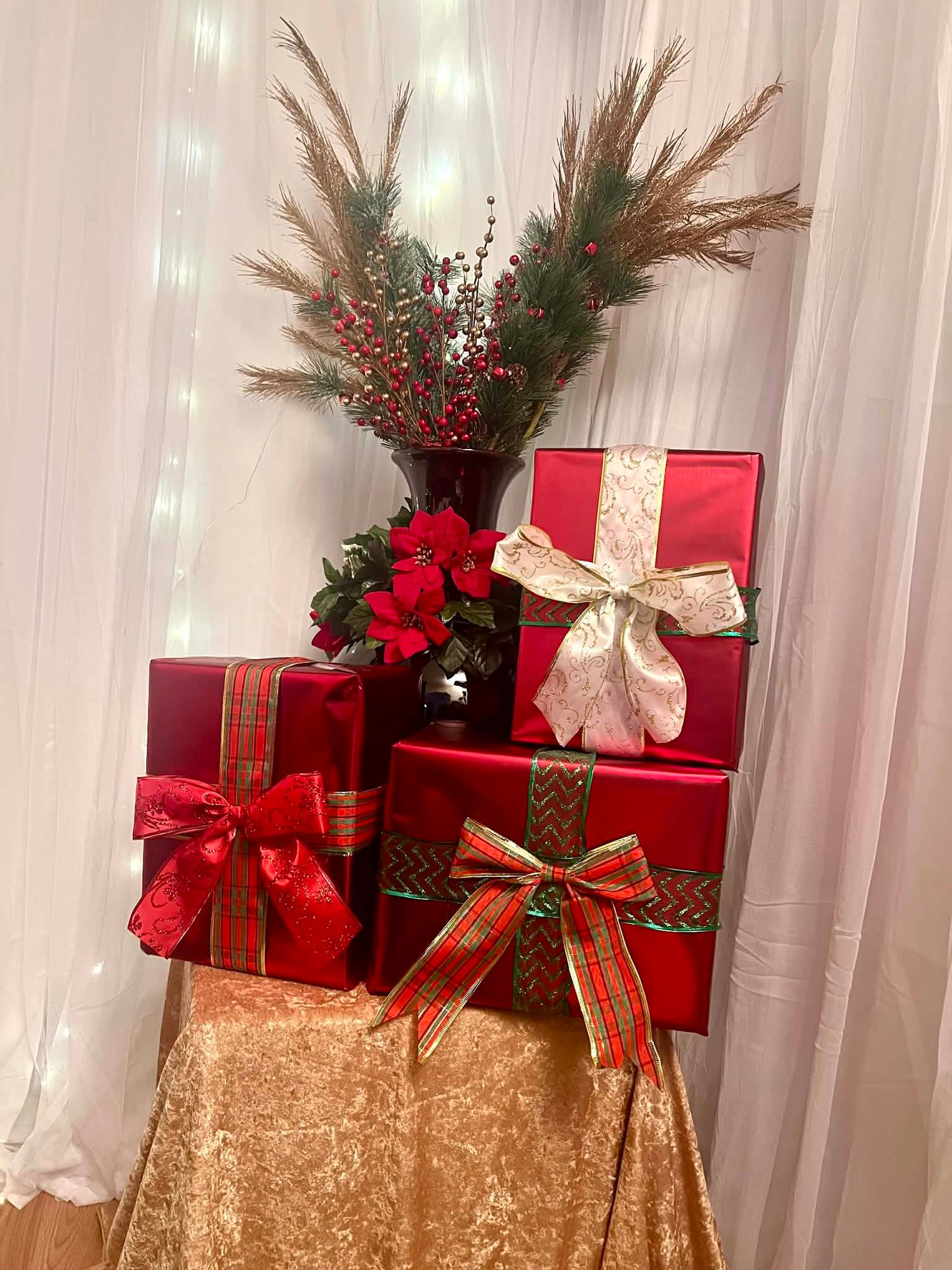 Red gift boxes with festive bows arranged near a vase with poinsettias and greenery against white drape backdrop.
