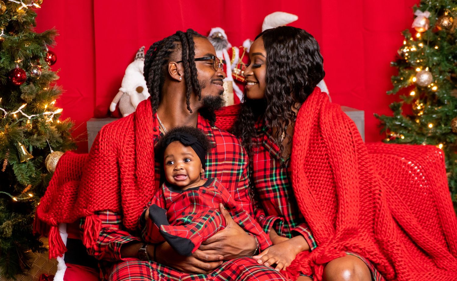 Happy family in red plaid pajamas sharing a kiss over smiling baby at Christmas.