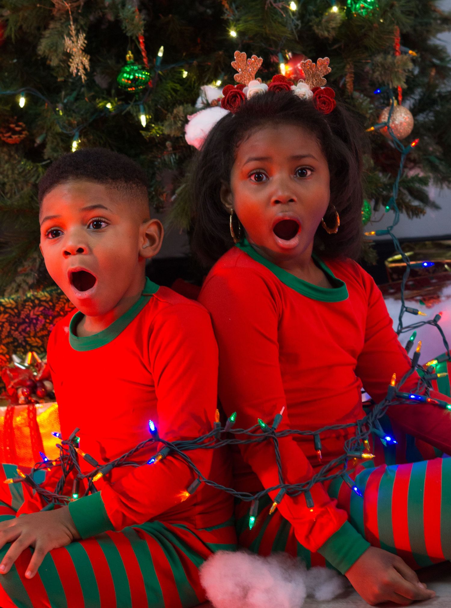 Two young children in red and green Christmas pajamas sit by tree with lights, mouths open in surprise.