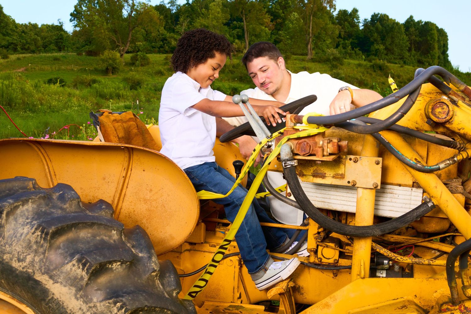 Two people sit on a yellow bulldozer in a green field, with a child holding the steering wheel.