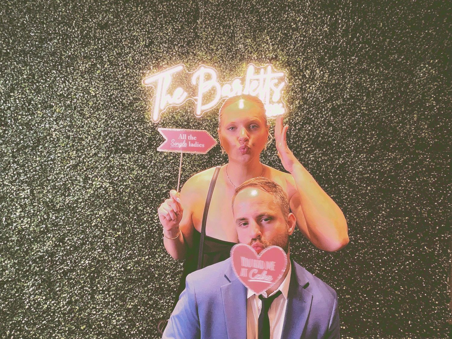 Couple posing at a wedding photo booth with neon sign and fun props against a dark floral wall.
