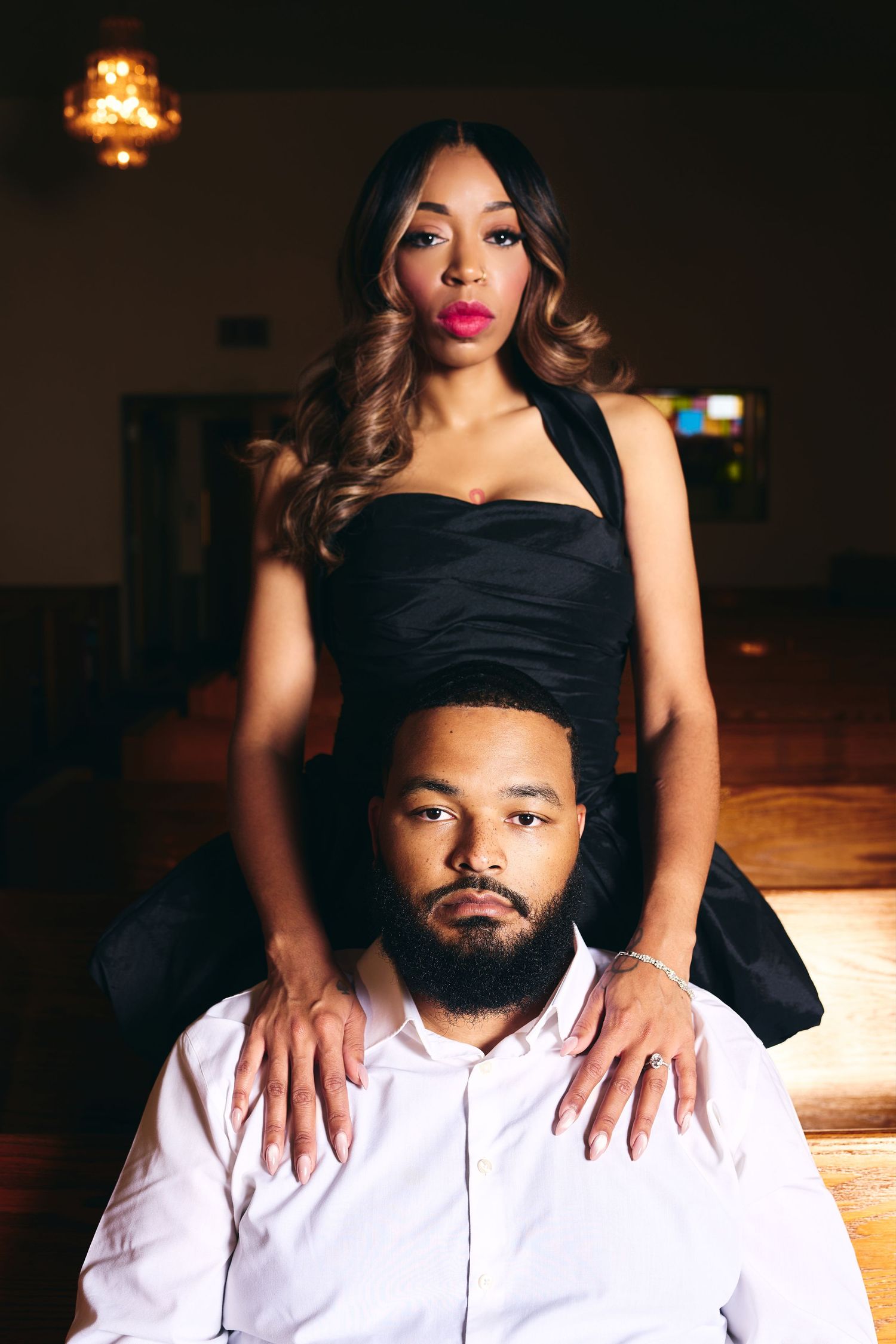 A stylish couple poses in a dimly lit venue, woman in black dress standing behind seated man in white shirt.