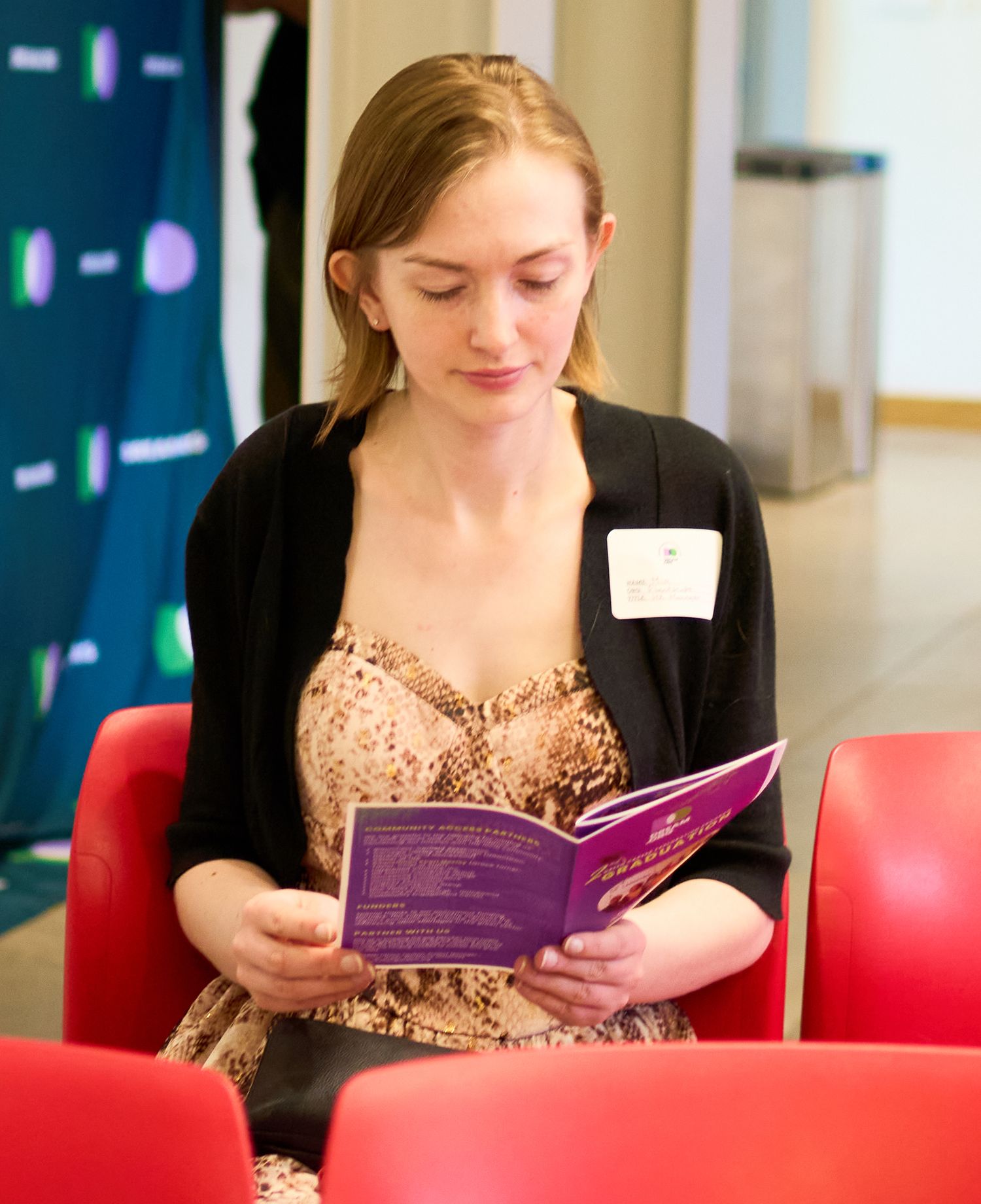 Young woman in floral dress reading a purple brochure at a professional event, wearing a name badge.