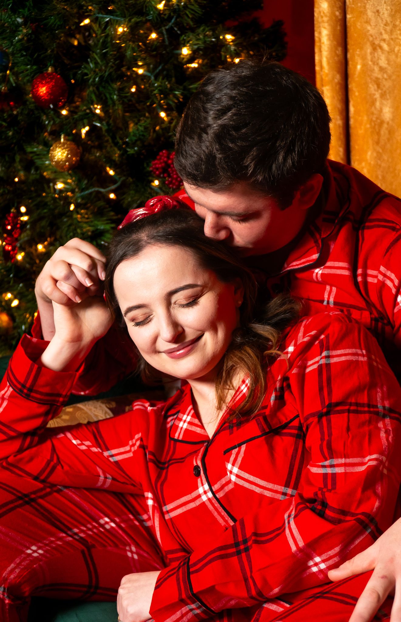 Couple in matching red plaid Christmas pajamas share a romantic moment beside a lit Christmas tree.
