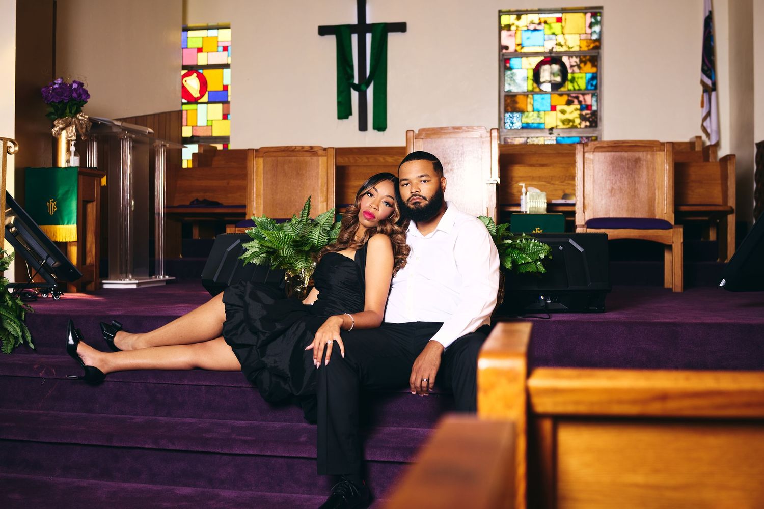 Couple posing elegantly in a church with purple pews, stained glass windows, and a green cross backdrop.