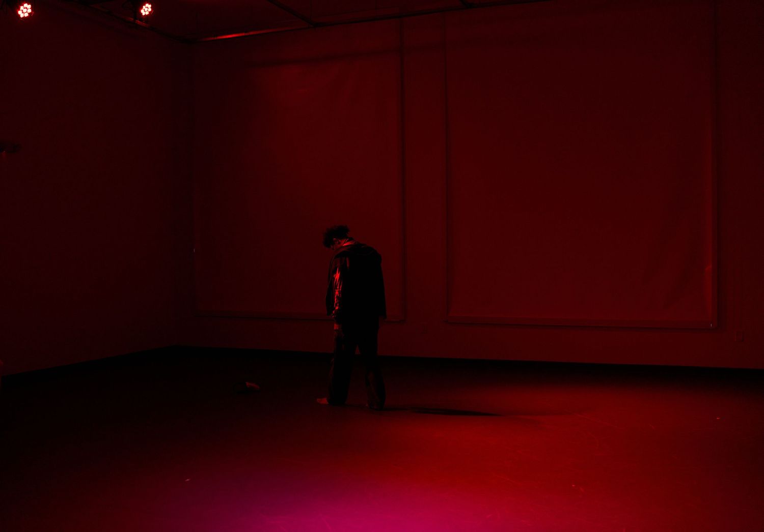 A lone figure stands in a dark room bathed in dramatic red lighting, creating a moody atmospheric scene.
