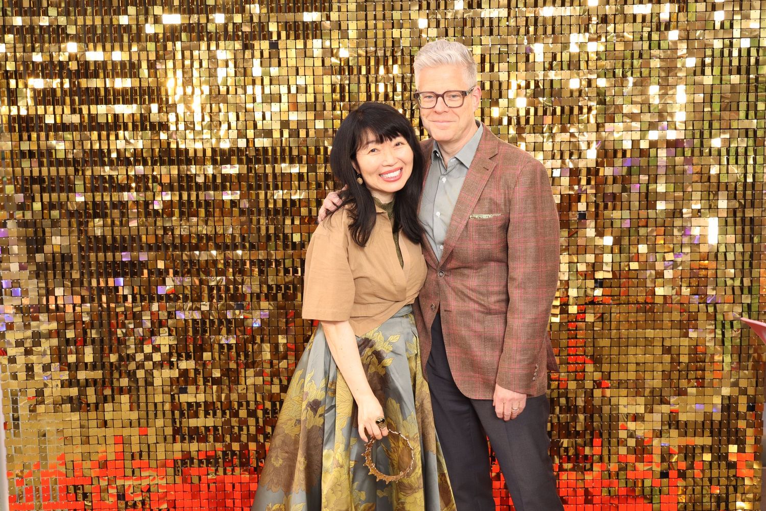 A woman and man pose together smiling in front of a gold sequin backdrop at a formal event.