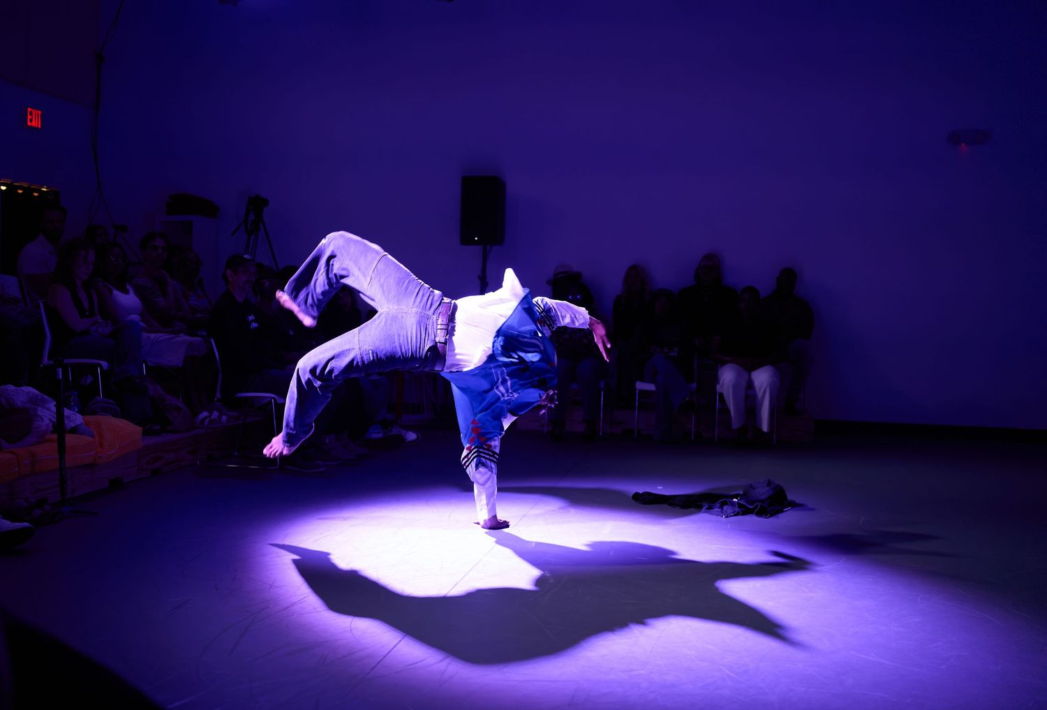 Breakdancer performing a one-handed freeze move under purple stage lighting with audience watching in background.