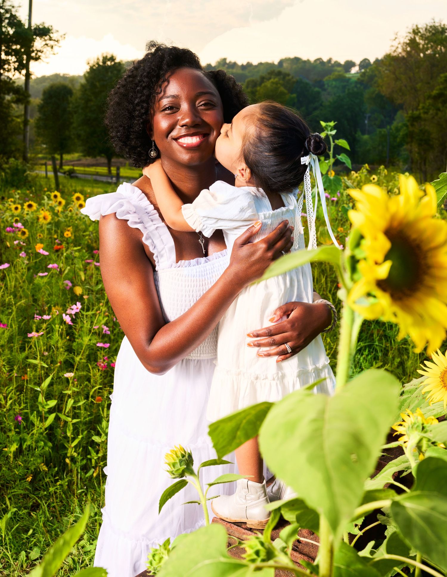 A smiling Black woman in a white dress holds a young girl kissing her cheek in a sunny sunflower field.