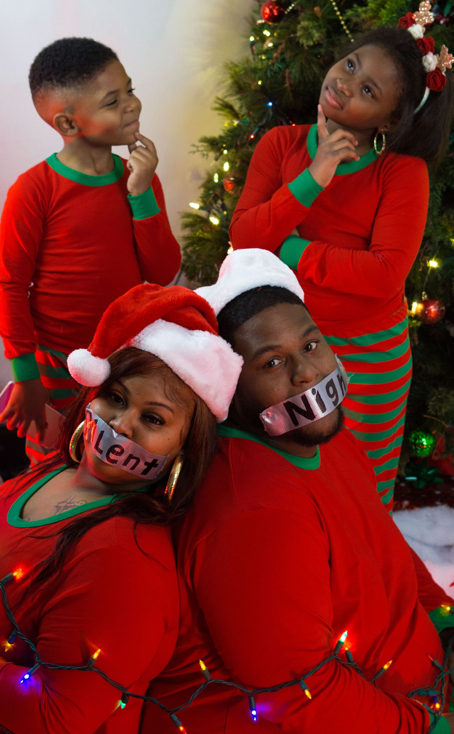 Family of four in red Christmas pajamas poses by tree, parents with tape on mouths, kids looking thoughtful.