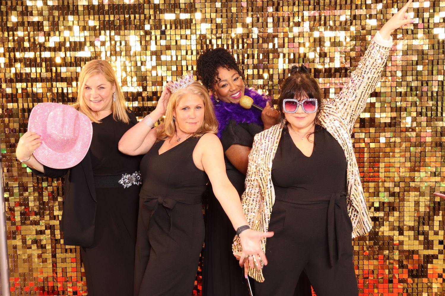 Four women posing in front of a gold sequin backdrop, wearing black outfits with fun accessories at a glamorous event.