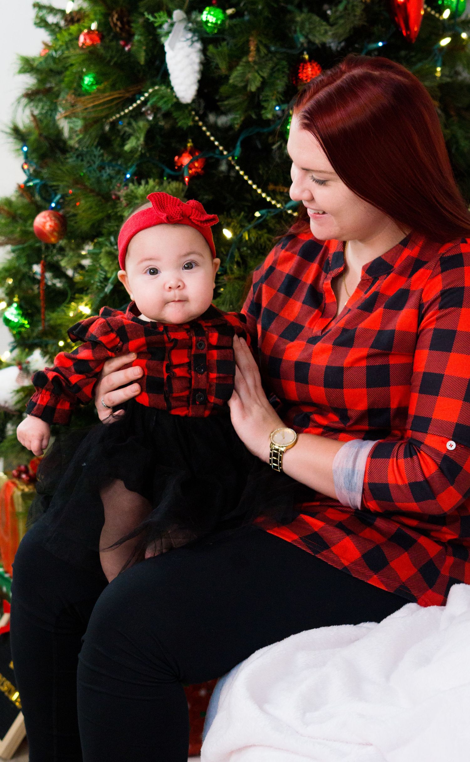 Mother and baby in matching red buffalo plaid outfits pose together in front of Christmas tree.