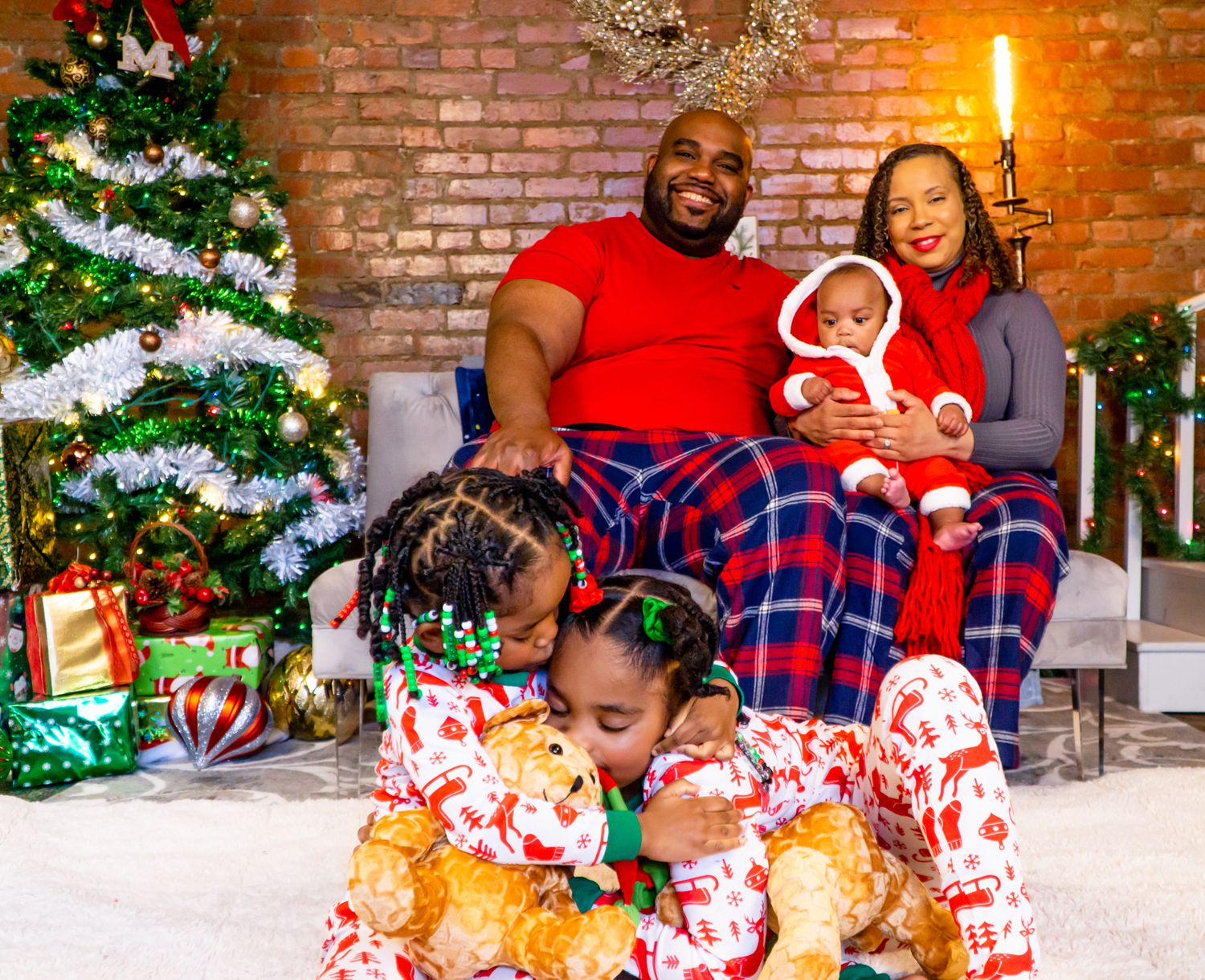 Family in matching Christmas pajamas poses near decorated tree with gifts, two girls hugging teddy bears in foreground.