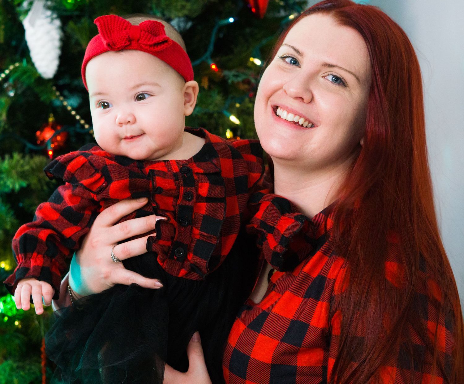 Mother and baby in matching red plaid outfits pose together in front of a decorated Christmas tree.