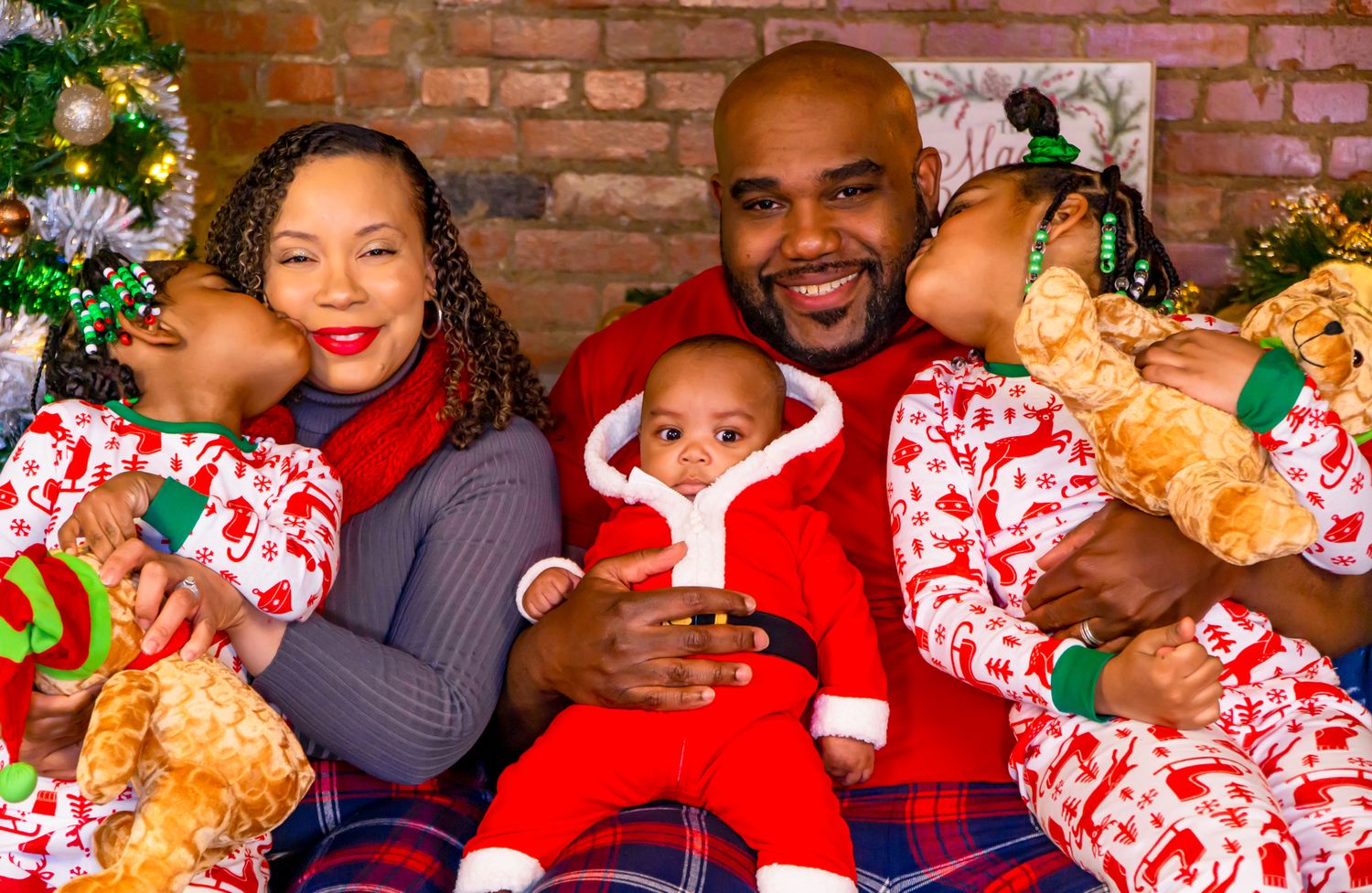 Happy family in Christmas pajamas cuddles together, parents holding children and baby in red Santa outfit.