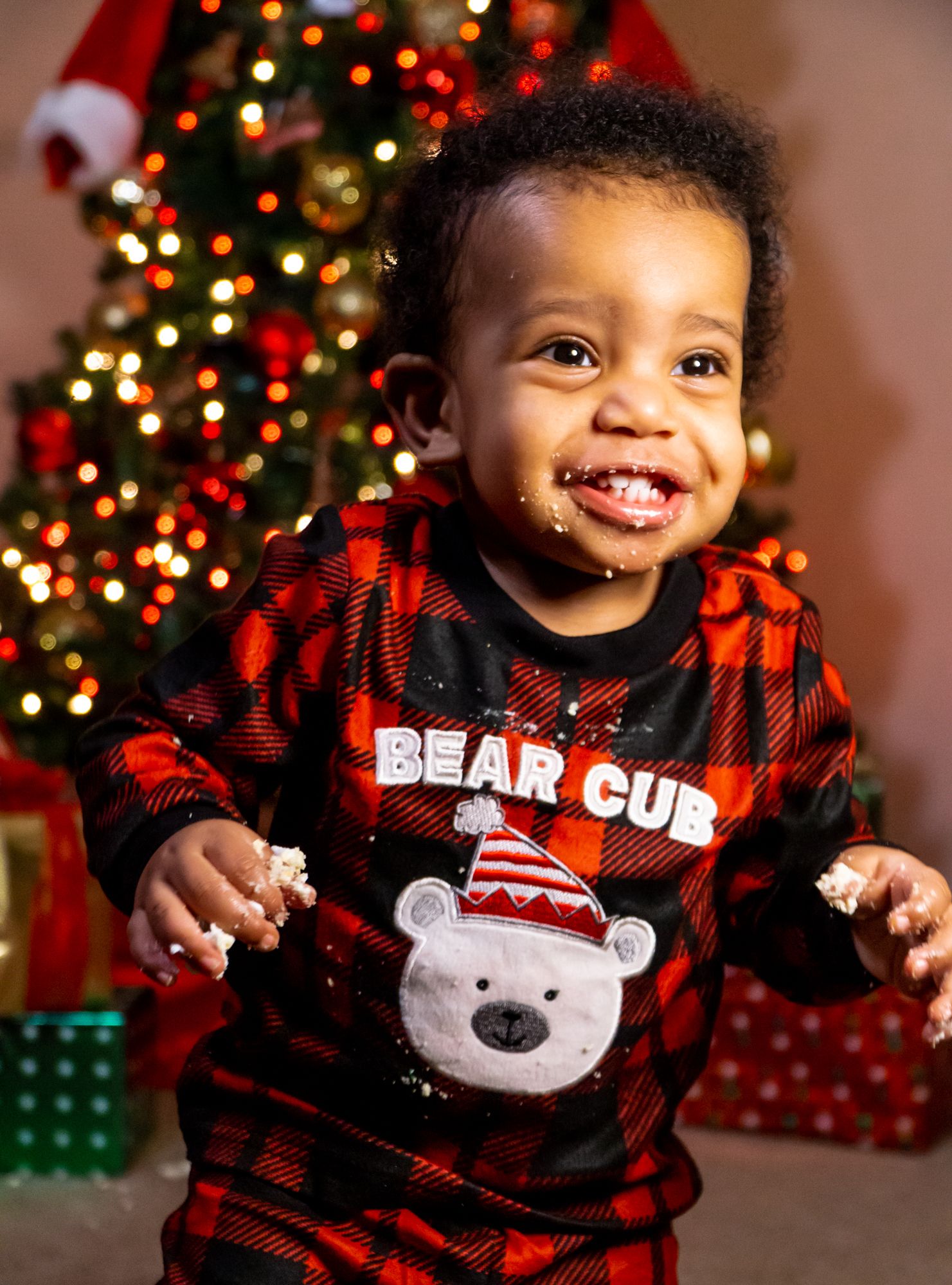 Adorable smiling toddler in red plaid Bear Cub pajamas standing by Christmas tree.